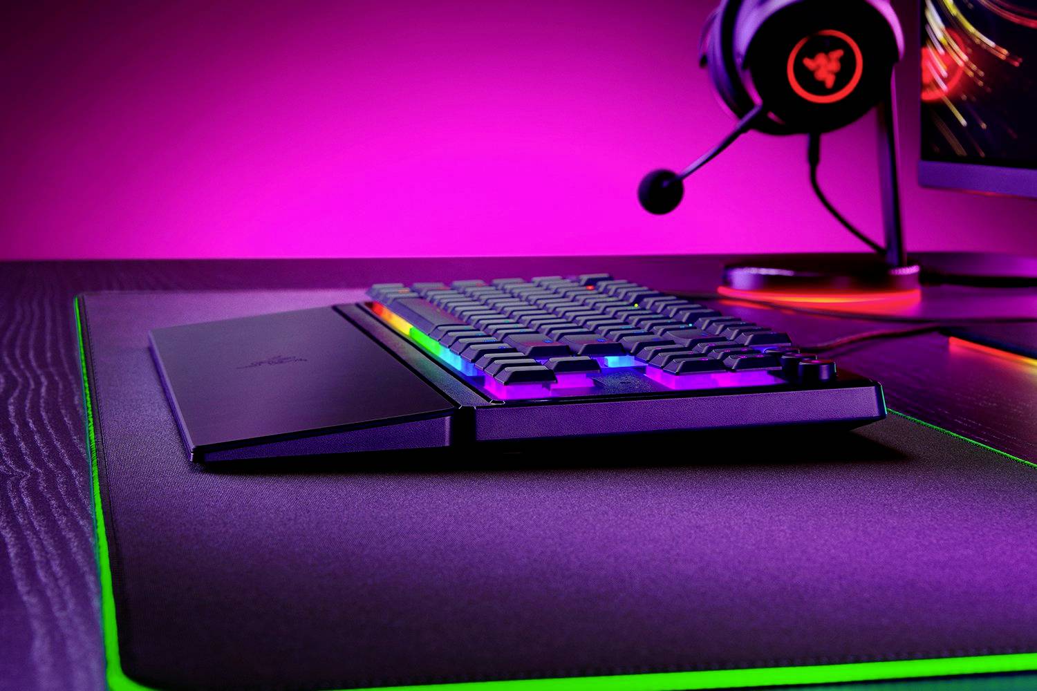 Clavier avec éclairage RVB sur un bureau, à côté d'un casque gaming lumineux et d'un écran avec un rétroéclairage violet.