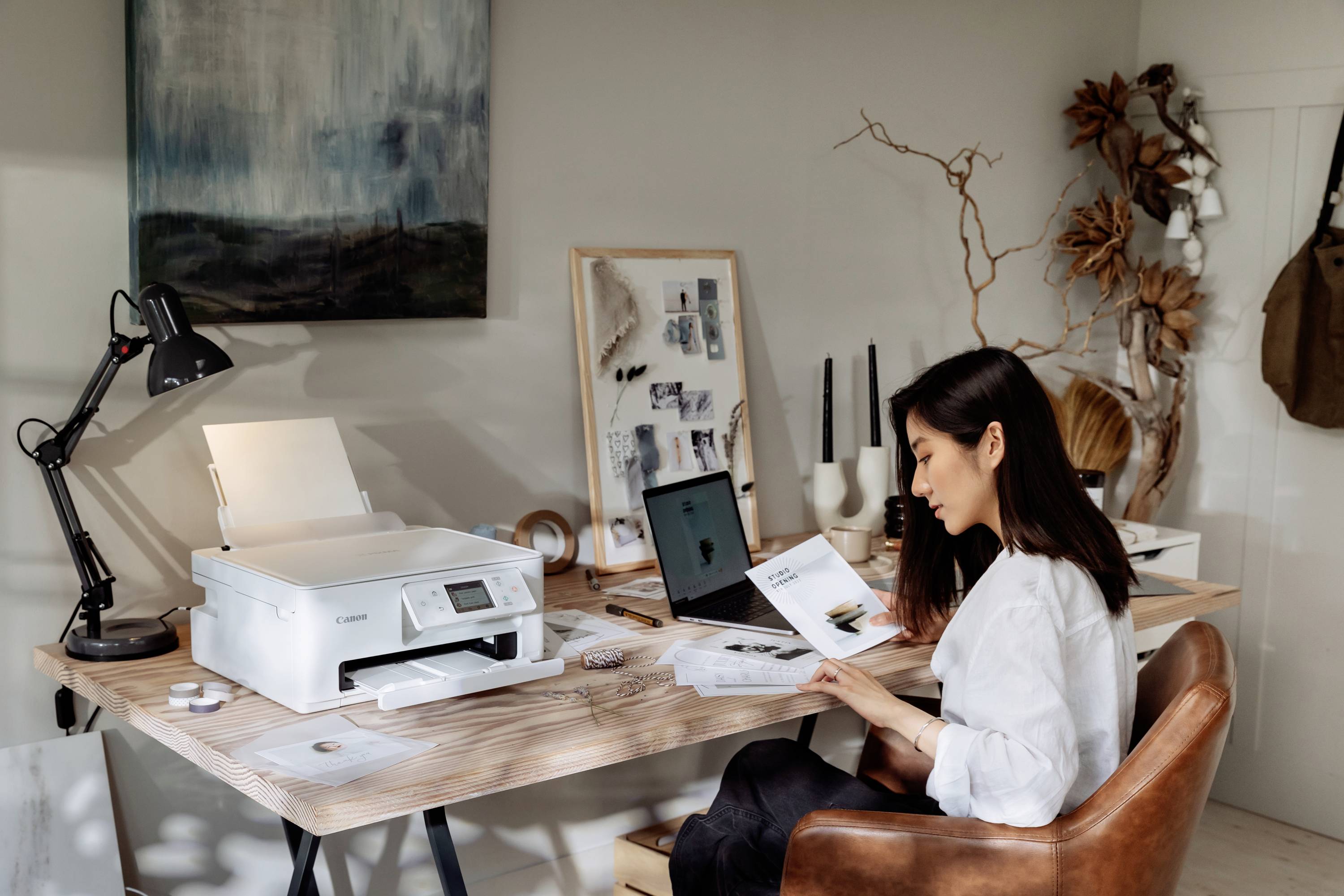 Une femme travaille à un bureau en bois avec une imprimante et un ordinateur portable. Elle lit attentivement un document.