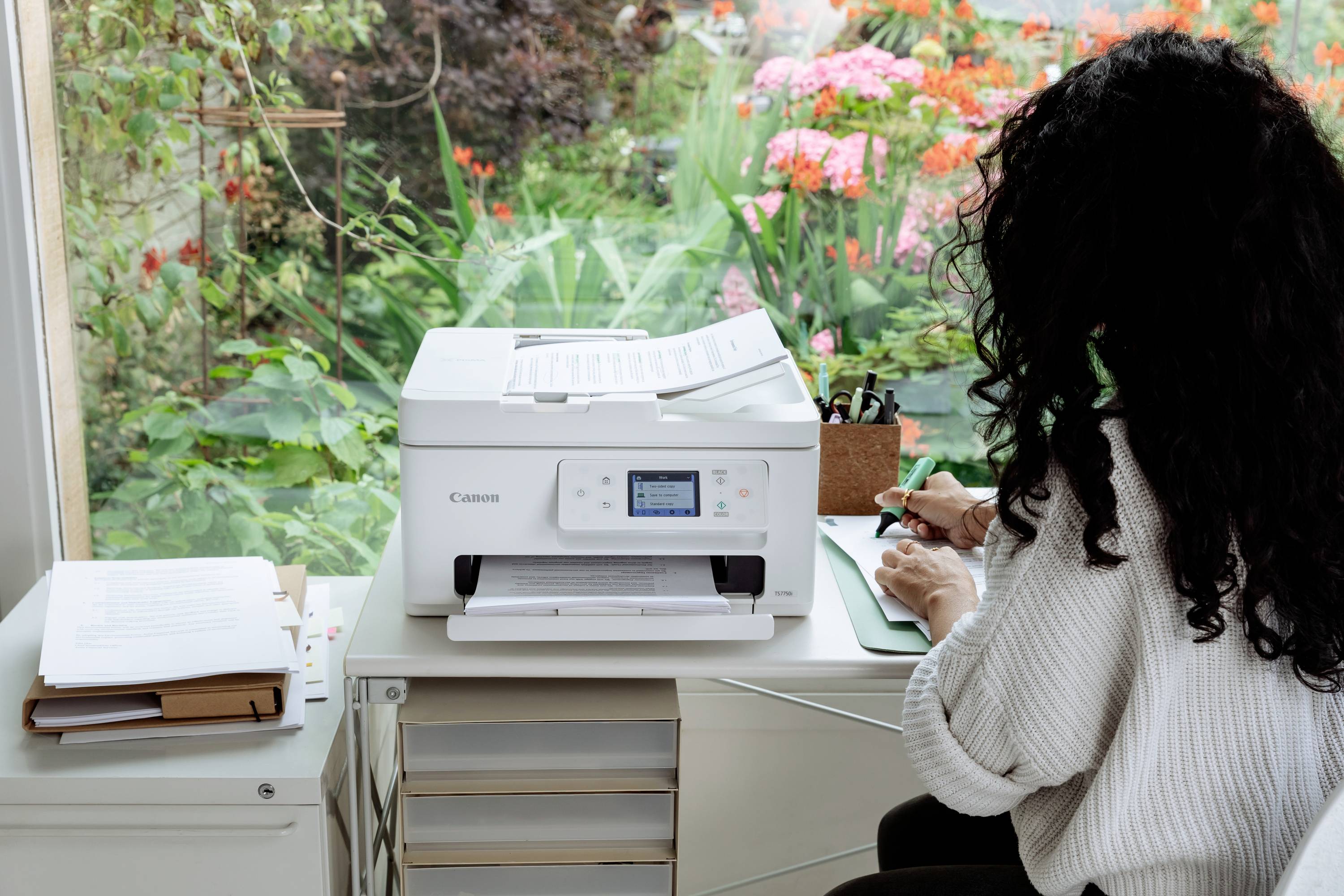 Une personne est assise à un bureau près d'une imprimante, écrit sur un bloc-notes. Un jardin en fleurs est visible en arrière-plan.