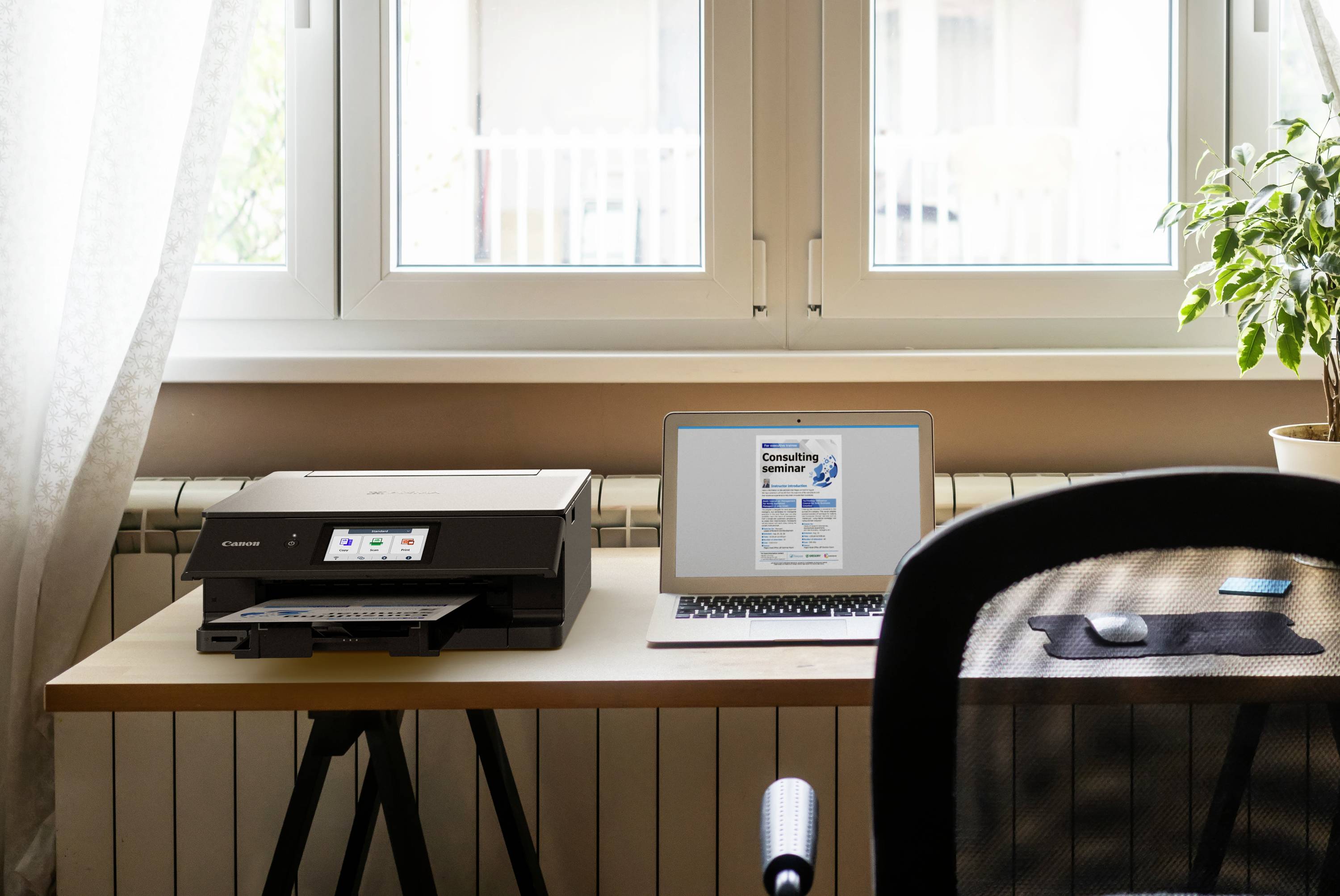 Poste de travail avec un ordinateur portable, une imprimante et une plante en pot sur une table près d'une fenêtre. L'ordinateur portable affiche un site web ouvert.