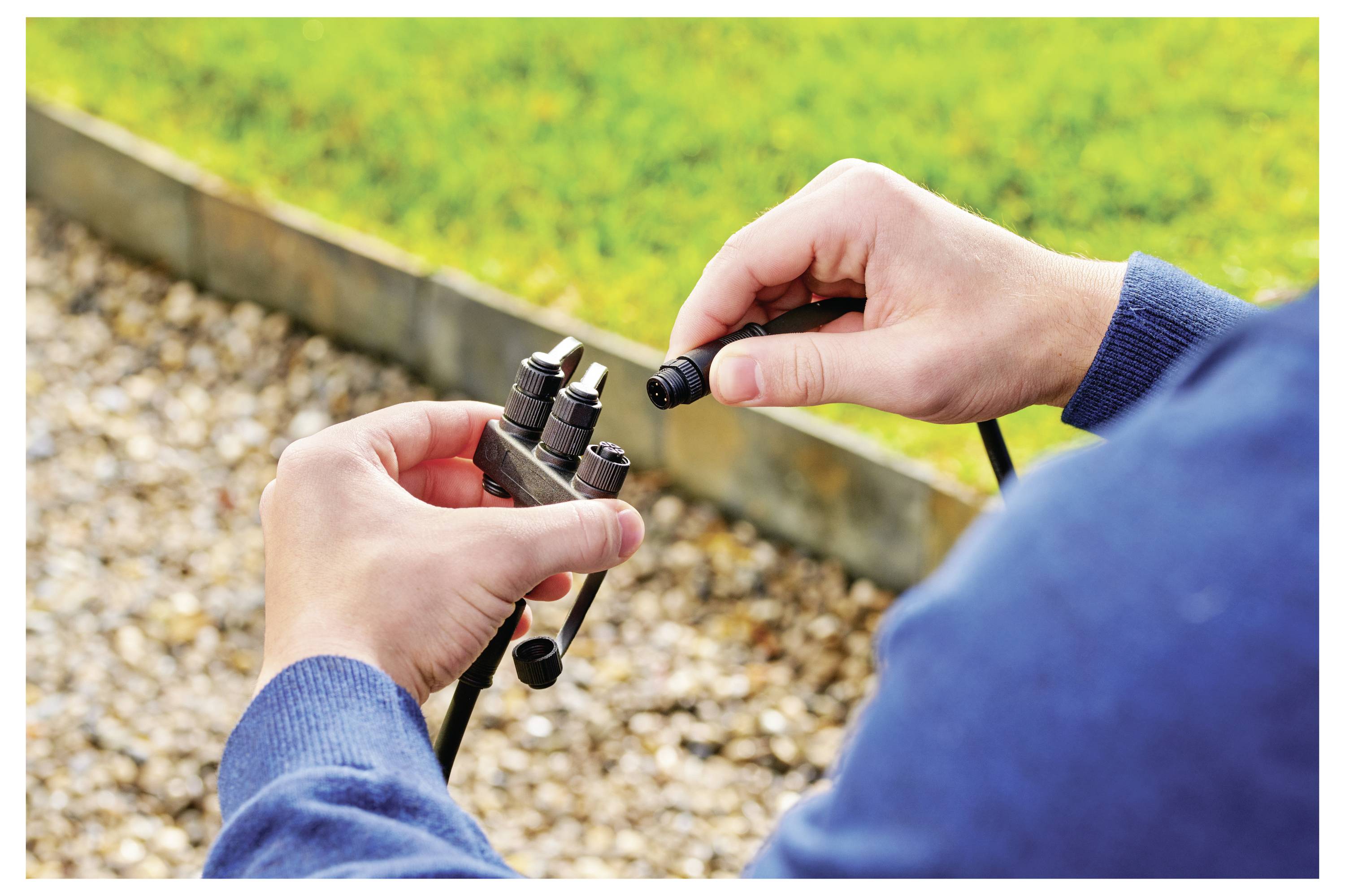 Personne portant un sweat-shirt bleu assemblant ou réparant un système d'irrigation, tenant des raccords et des tuyaux dans une zone de jardin rocheuse.