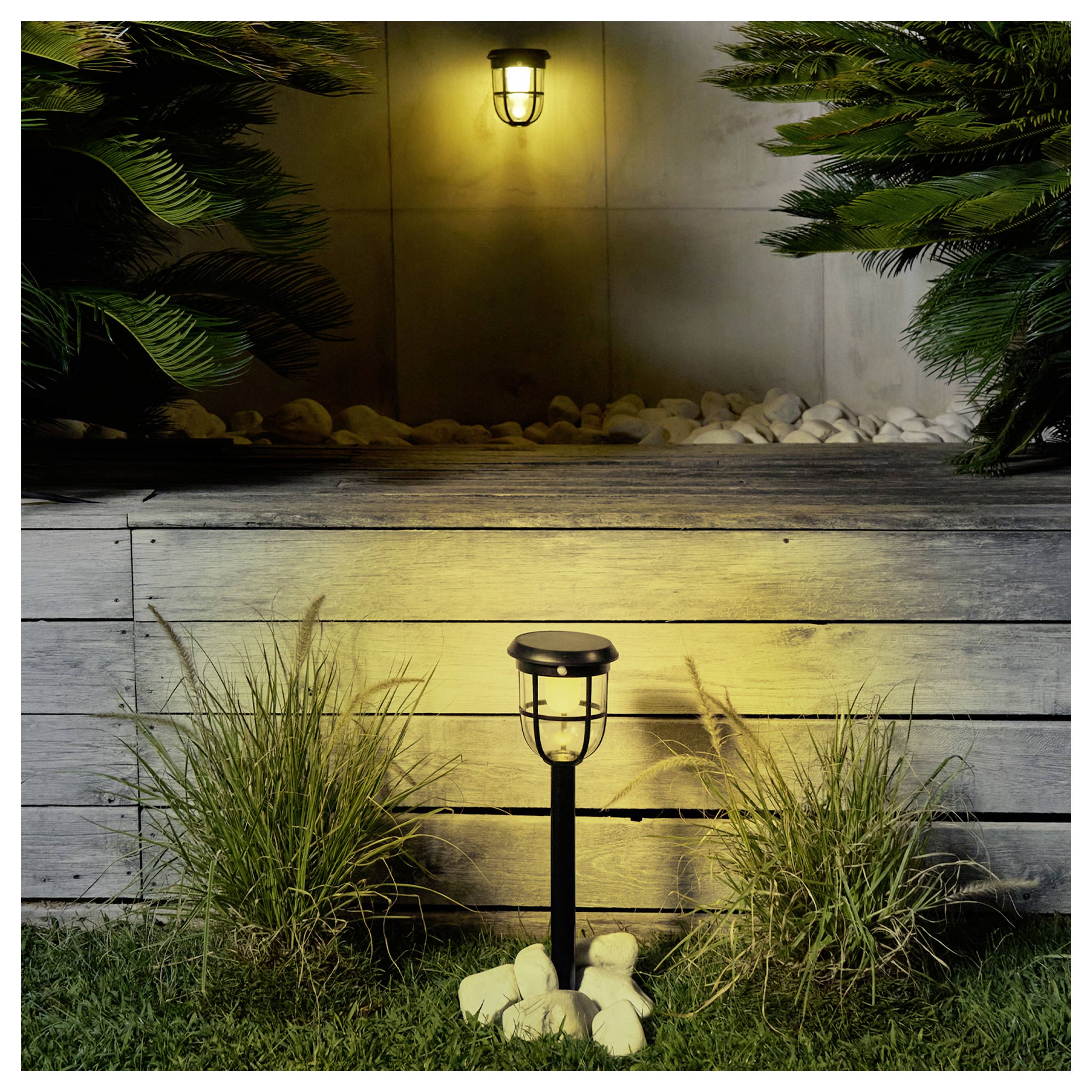 Poteau de lampadaire de jardin extérieur devant un mur de jardinière surélevée, avec une lanterne brillante et des plantes décoratives la nuit.
