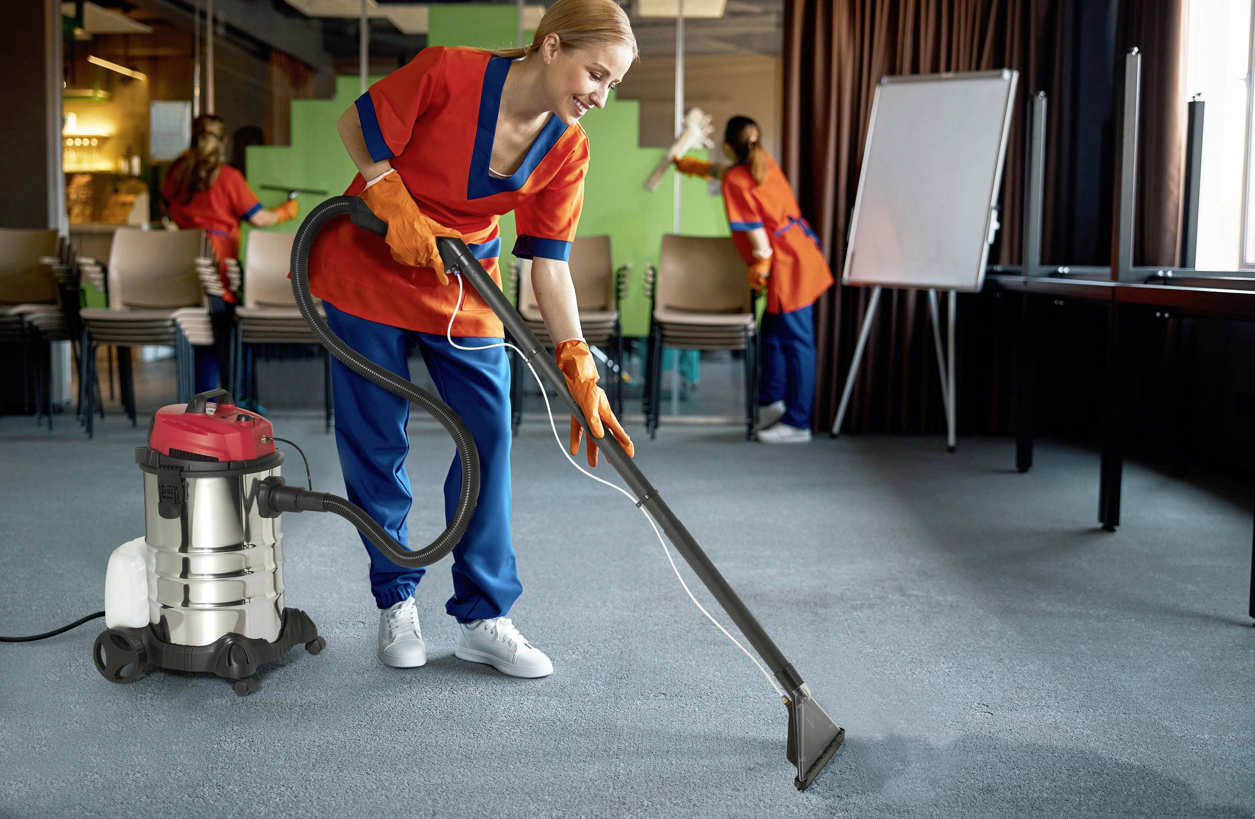 Une femme en tenue de travail aspire le sol moquetté d'une salle de conférence à l'aide d'un aspirateur industriel. À l'arrière-plan, une autre personne nettoie la pièce.