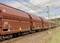 Un long train de marchandises avec des wagons bruns circule sur une voie ferrée sous un système de lignes électriques par une journée ensoleillée.