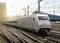 Un train à grande vitesse moderne roule sur des rails en passant devant une gare au coucher du soleil. L'environnement montre des caténaires.