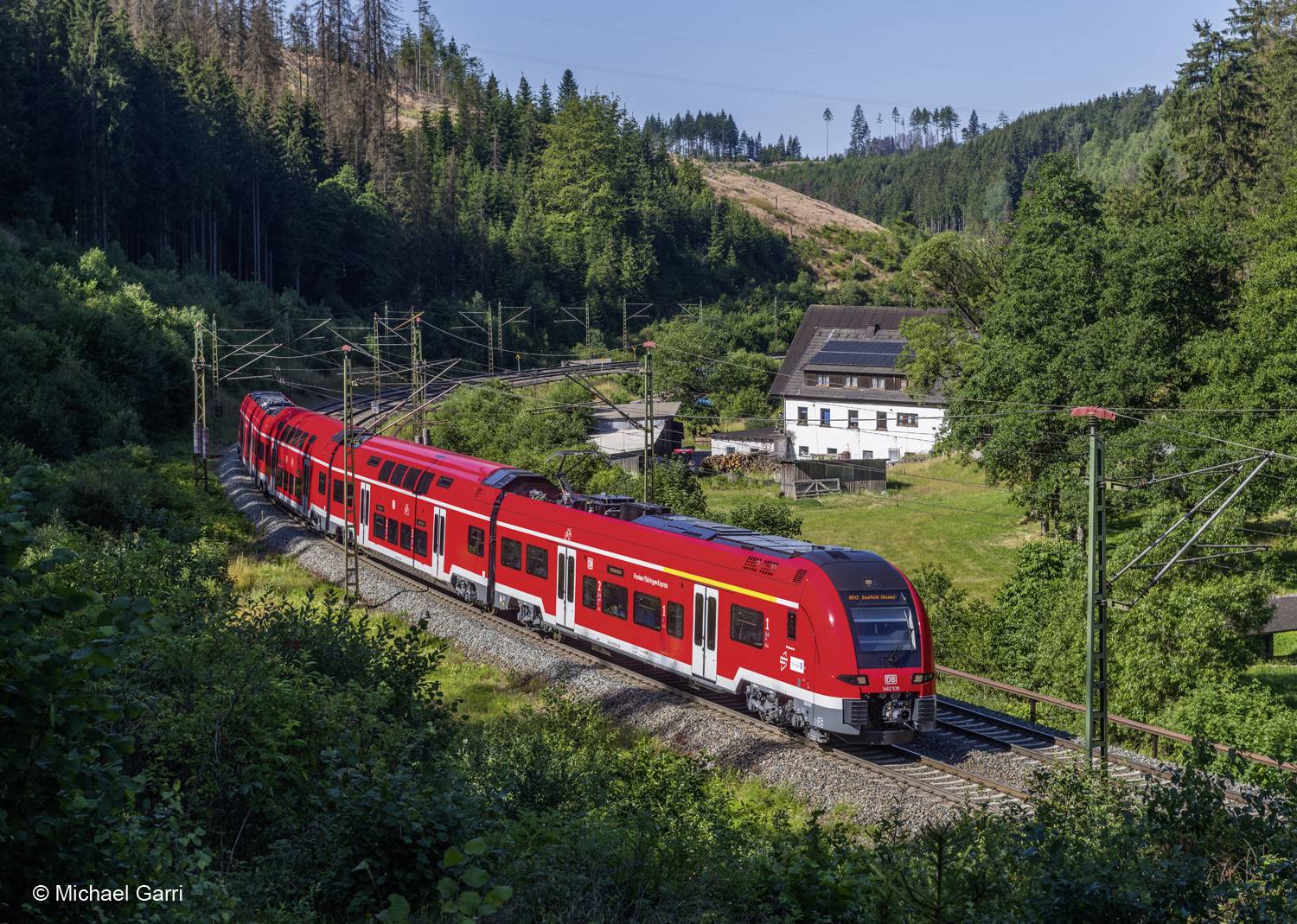 Un train régional rouge circule pendant la journée à travers un paysage boisé. Un bâtiment blanc est visible en arrière-plan.