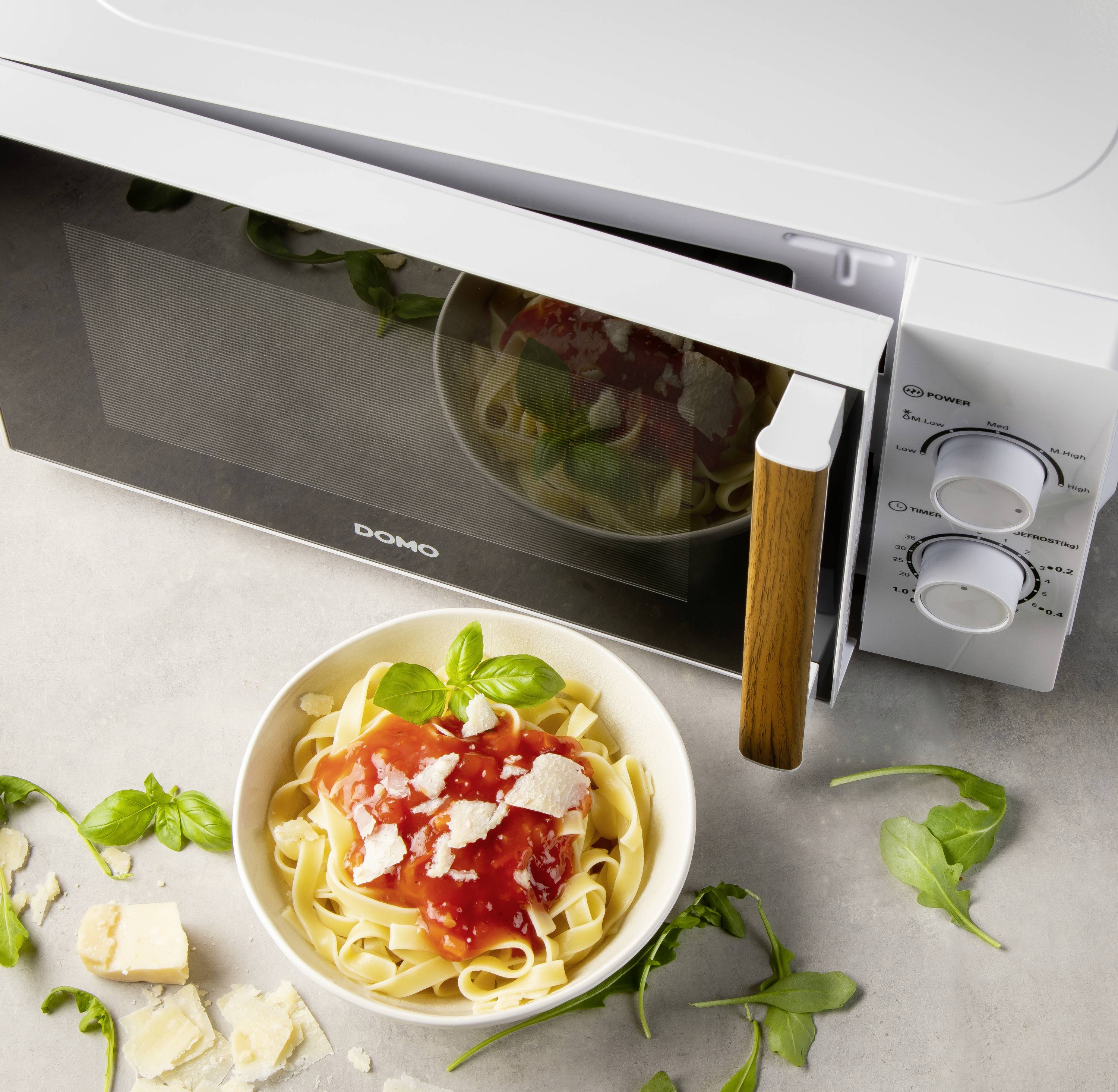Une assiette de tagliatelles, sauce tomate et basilic est posée à côté d'un four à micro-ondes blanc ouvert. Des morceaux de parmesan sont dispersés sur la table.