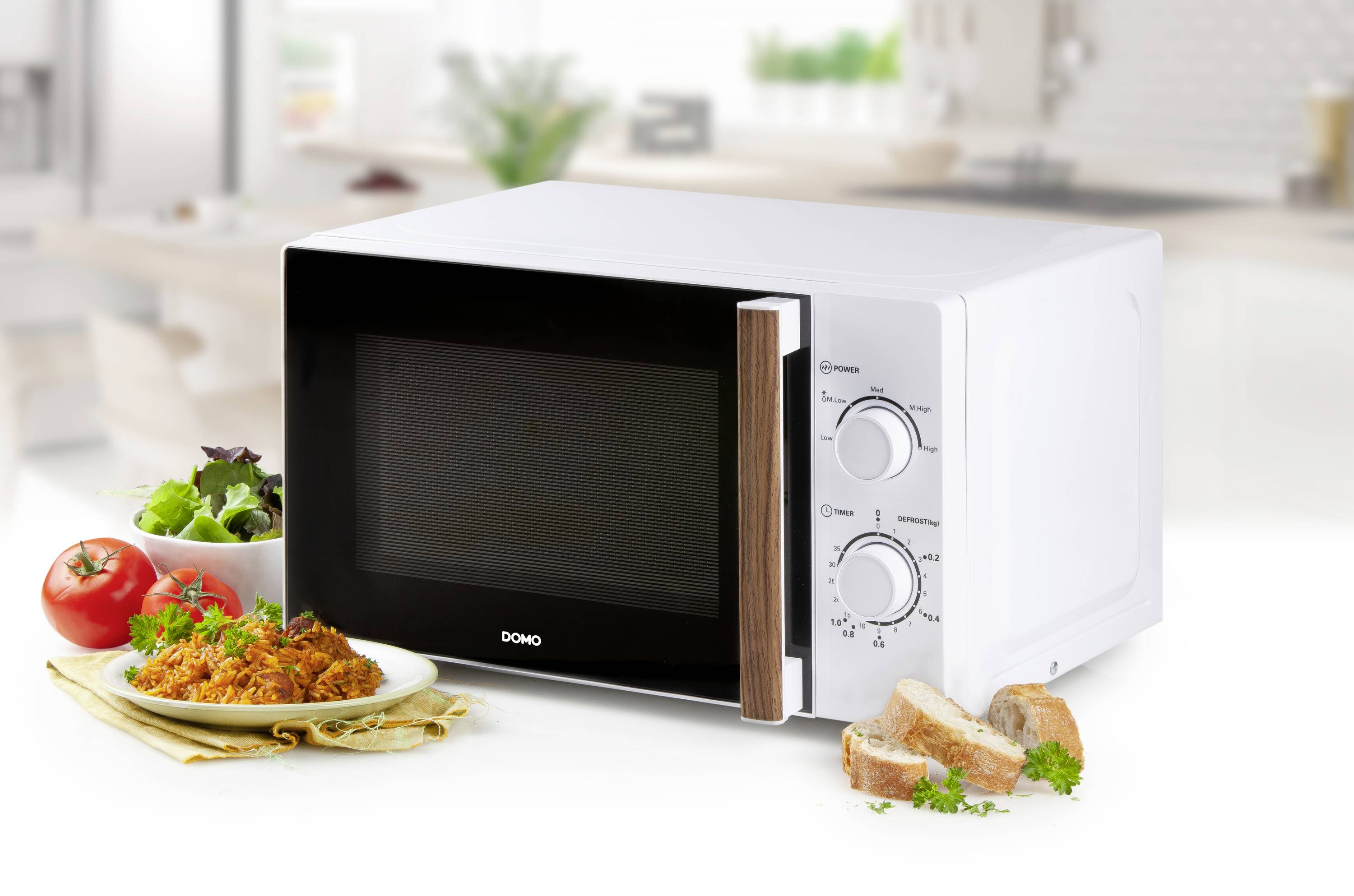 Four blanche posée sur une table de cuisine, à côté d'une assiette contenant un plat de riz, une salade, des tomates et du pain. L'appareil est équipé de deux boutons rotatifs pour les réglages.