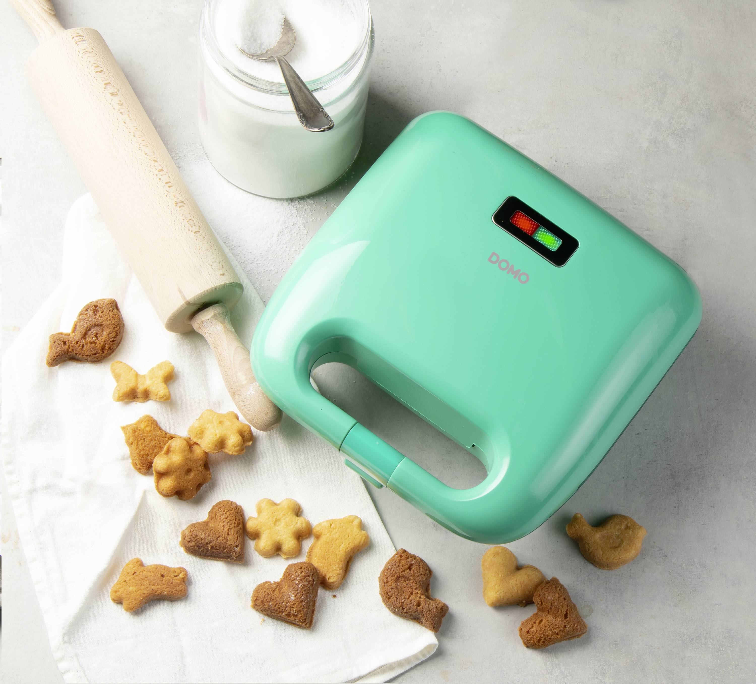 Un sandwich maker turquoise à côté de biscuits en forme d'animaux, d'un rouleau à pâtisserie et d'un bocal de sucre, posés sur un tissu sur un fond clair.