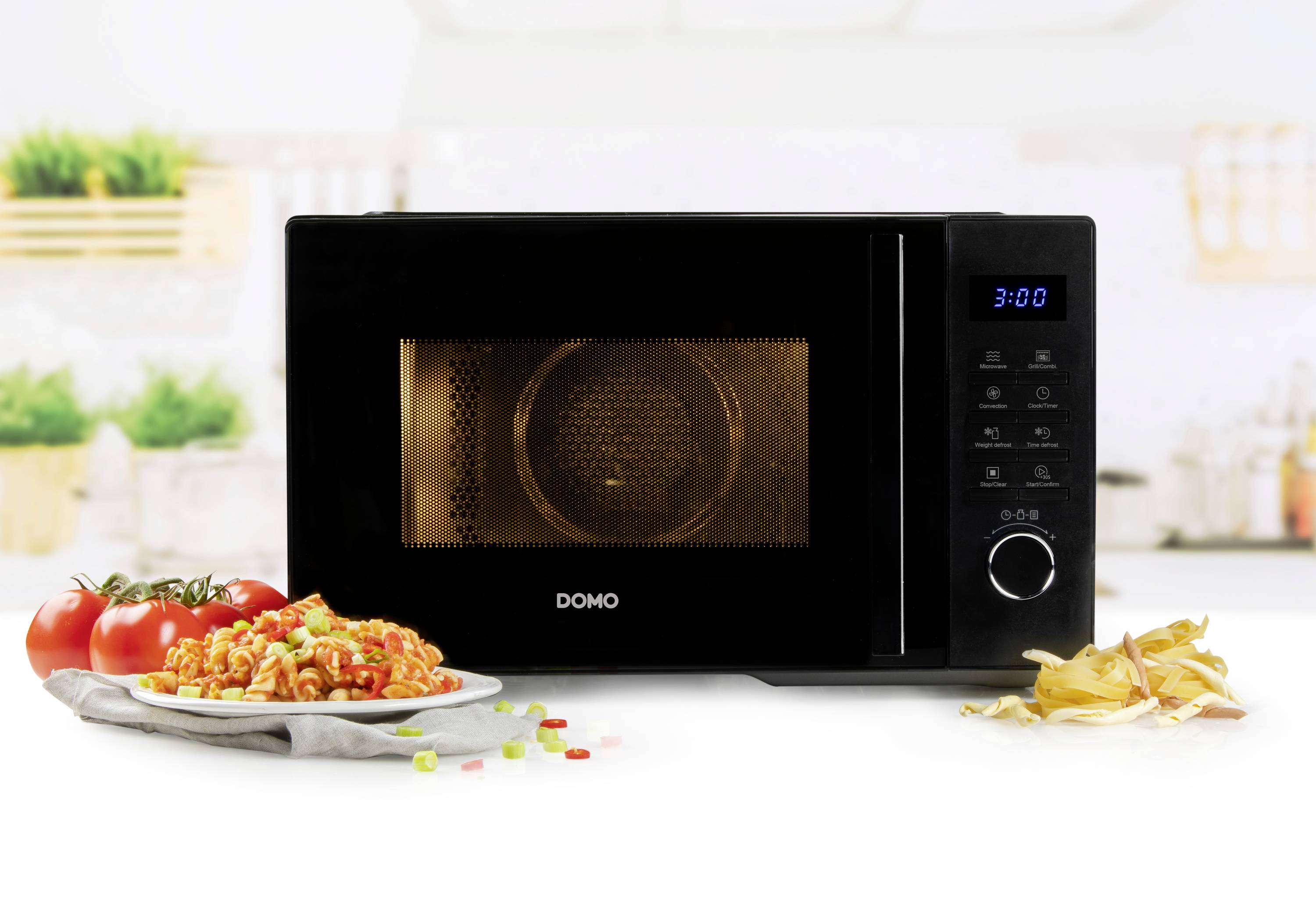 Four à micro-ondes noir sur un plan de travail de cuisine, à côté d'une assiette de pâtes et de tomates. En arrière-plan, une scène de cuisine floue.