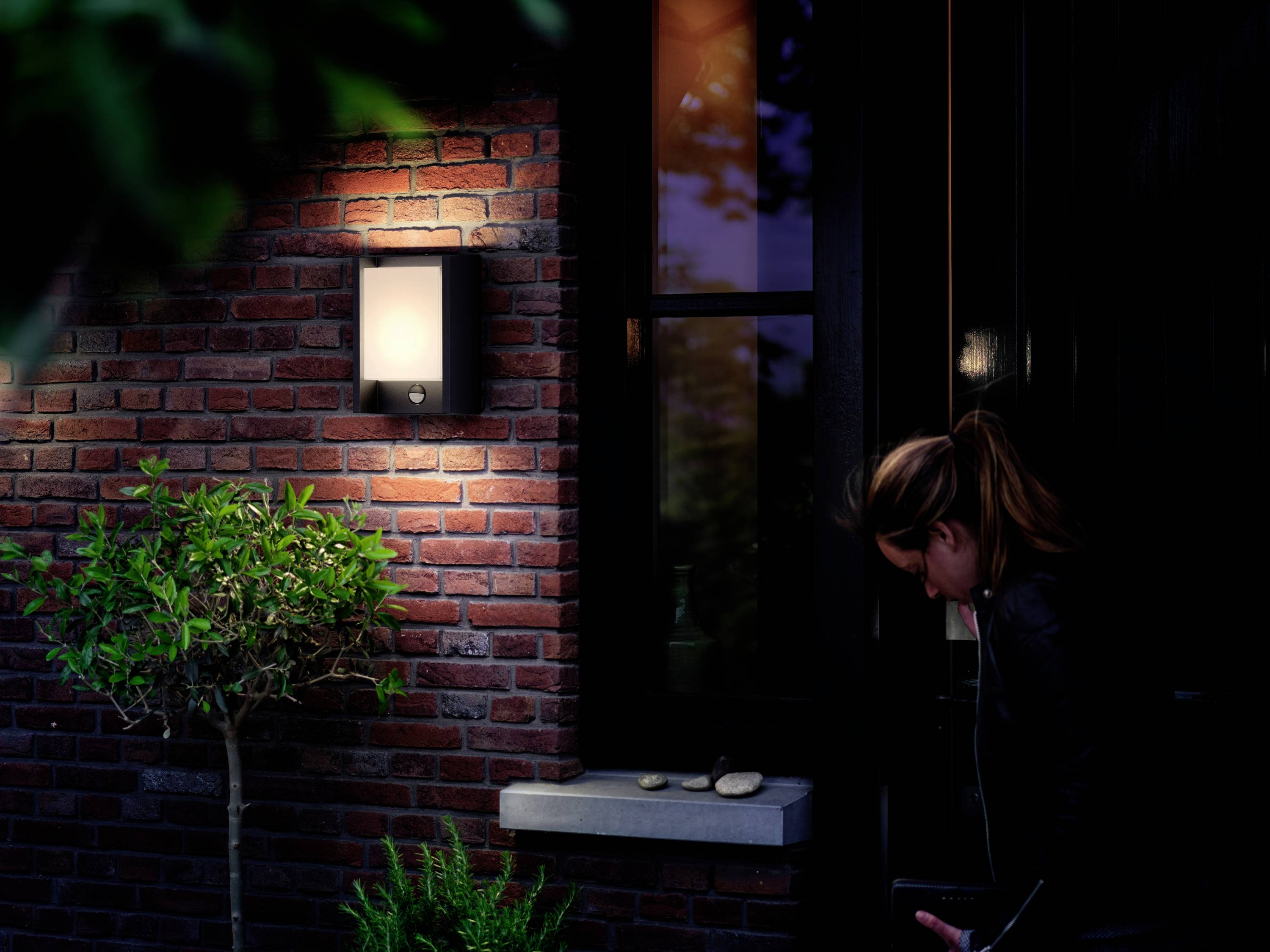 Une femme se tient devant une porte d'entrée sombre la nuit, éclairée par une lampe fixée au mur de briques. Un petit arbre se trouve à côté de la porte.