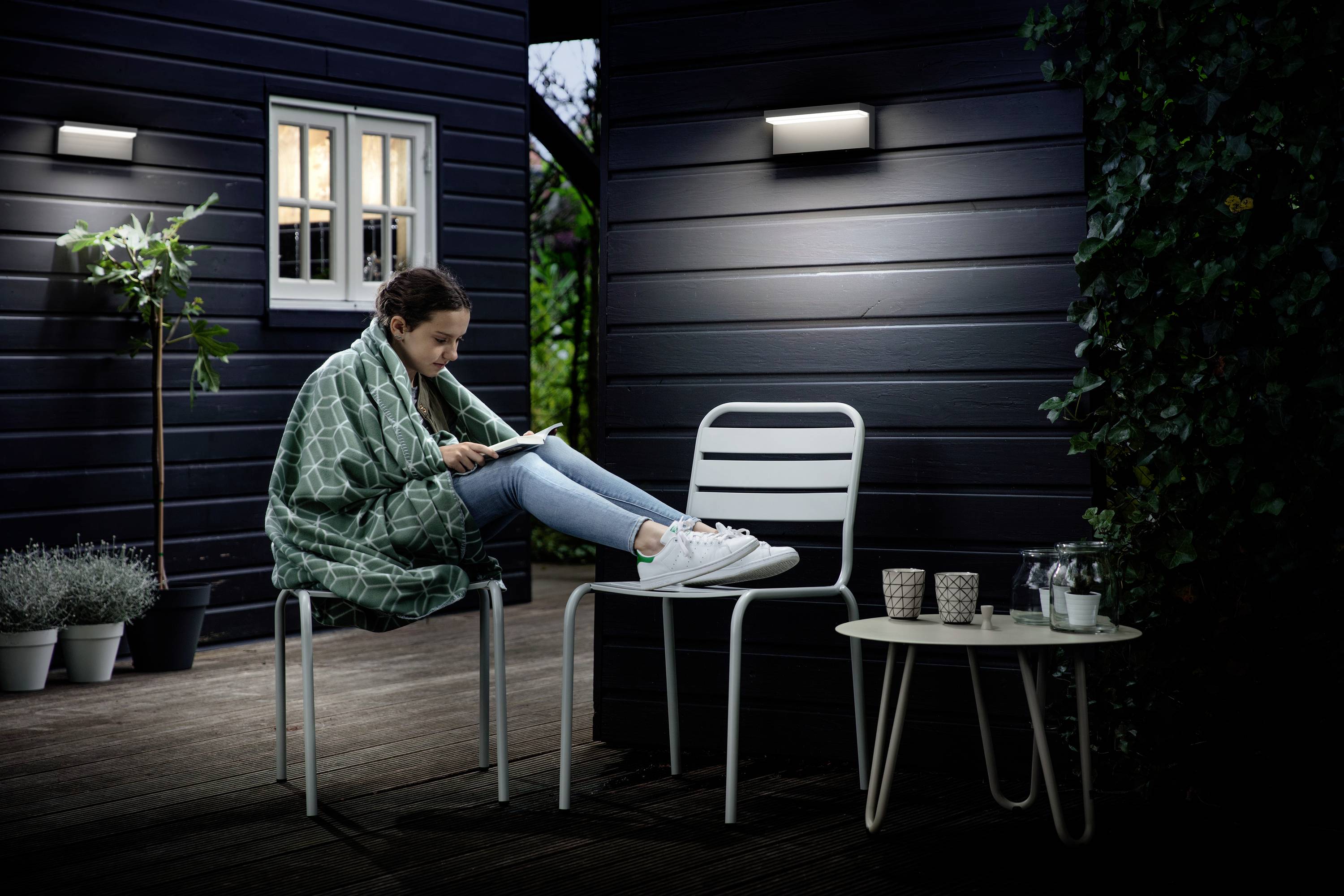 Une personne est assise, enveloppée dans une couverture sur une terrasse, en train de lire un livre. Le soir est tombé, et l'ambiance est doucement éclairée.