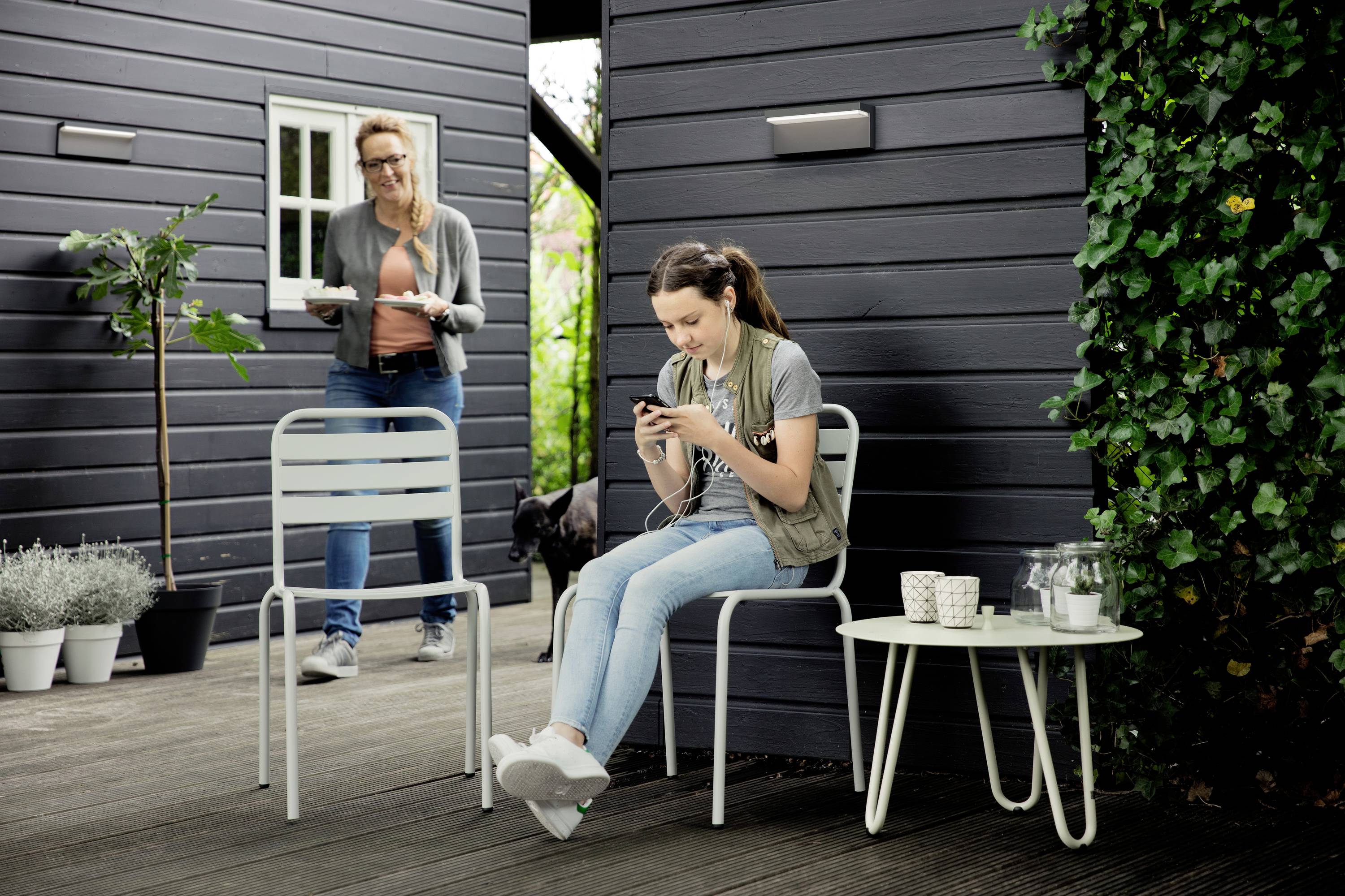 Une femme apporte des boissons et des biscuits sur un plateau, tandis qu'une jeune fille est assise sur une chaise et joue sur son téléphone portable. Un chien se trouve en arrière-plan.