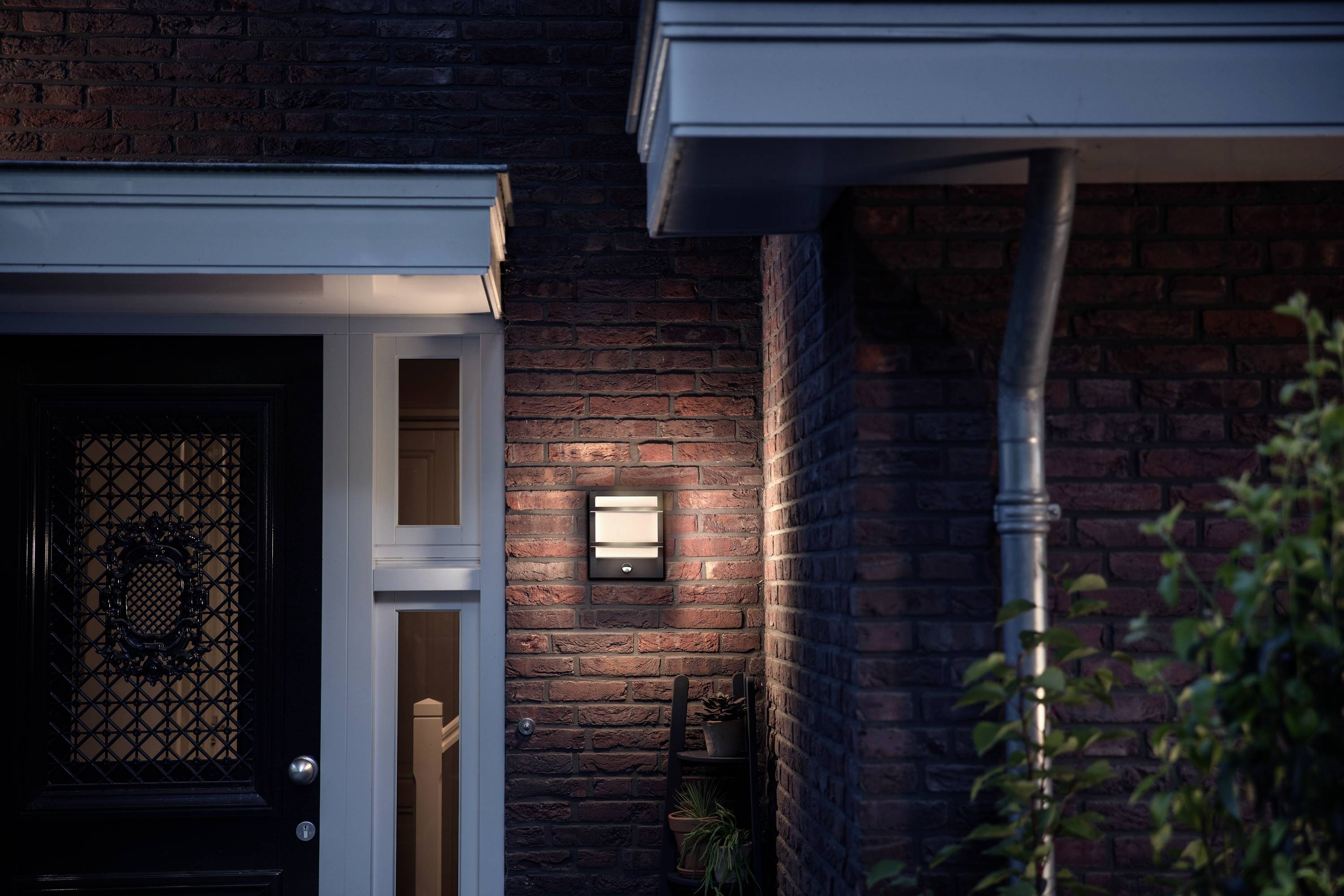 Une maison éclairée avec une façade en briques la nuit. Une lampe projette de la lumière sur le mur à côté d'une porte d'entrée noire.
