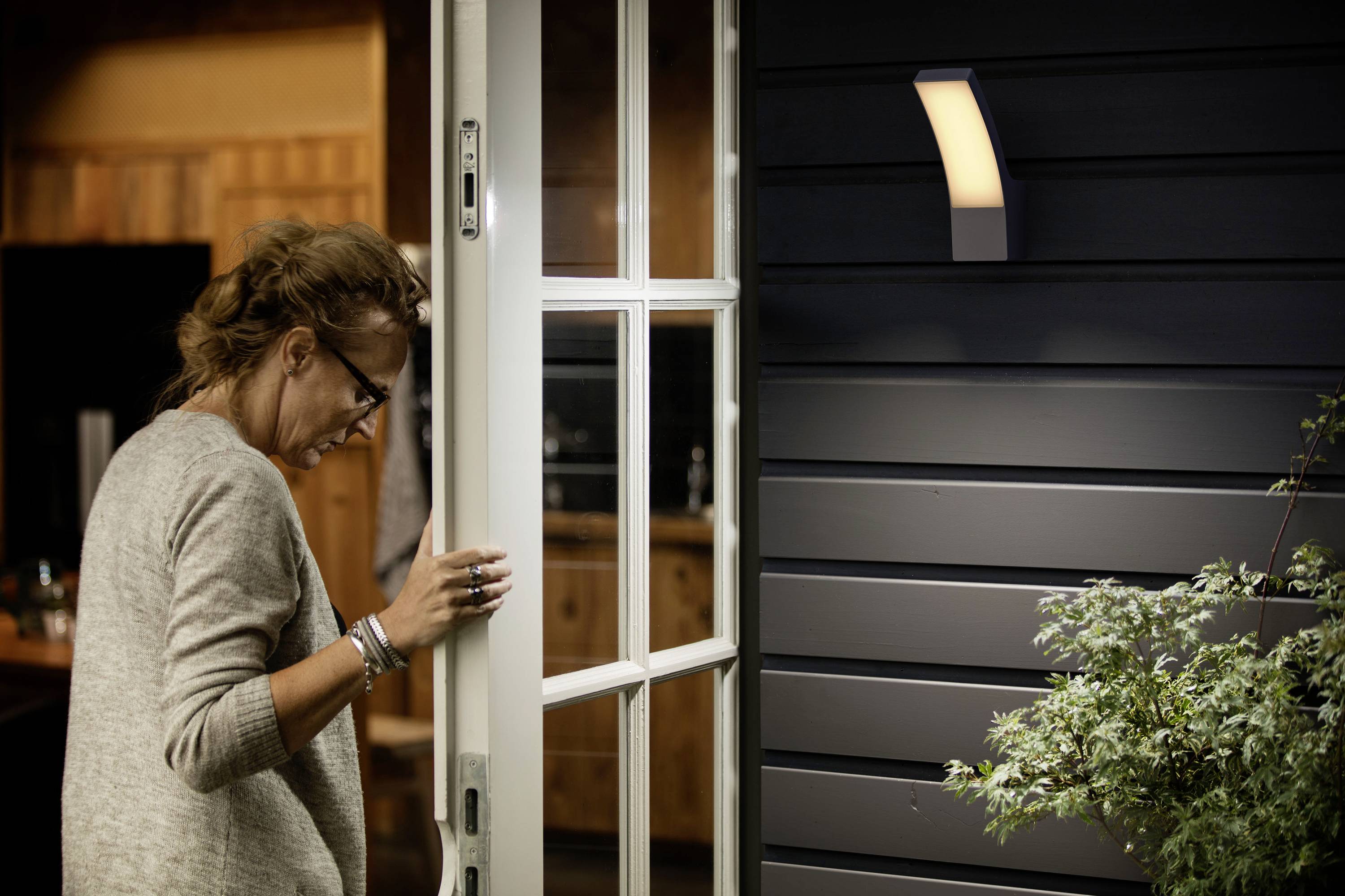 Une personne ouvre une porte vitrée vers l'extérieur. À l'arrière-plan, on aperçoit un intérieur en bois, tandis qu'une applique murale brille à l'extérieur.