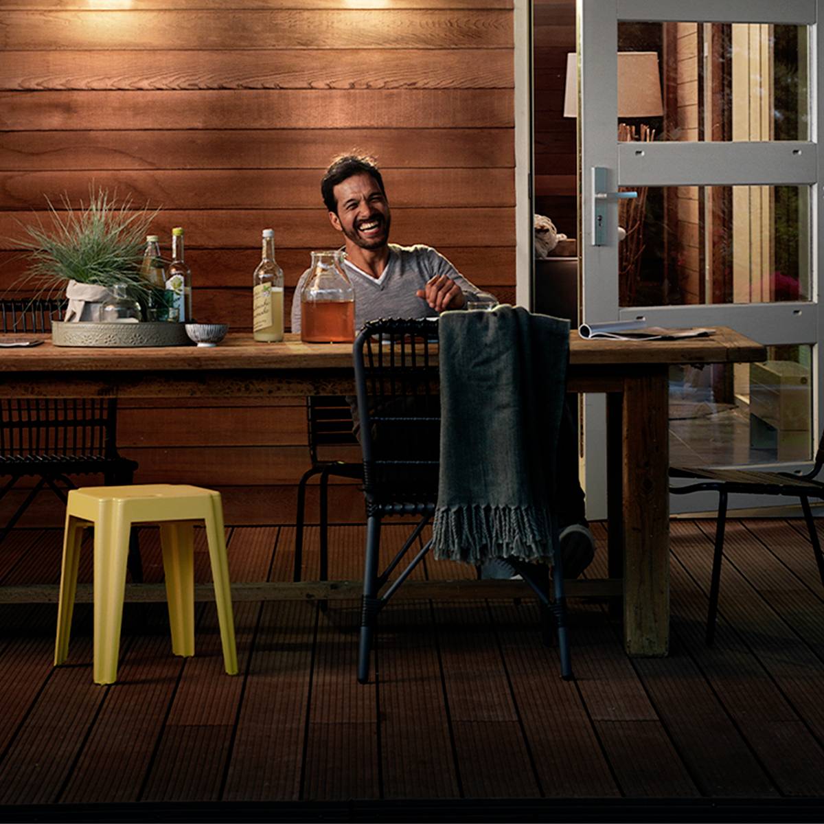 Une personne est assise à une table sur une terrasse, souriante, avec des bouteilles de boisson et des plantes en arrière-plan.