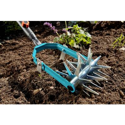 Un aérateur manuel bleu pour le sol, équipé de roues à pointes, est placé sur un sol fraîchement labouré. Des plantes vertes et des feuilles sont visibles en arrière-plan.