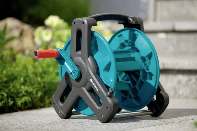 Un enrouleur de tuyau d'arrosage turquoise avec une poignée rouge est posé sur une terrasse à proximité de plantes vertes et d'une marche en pierre.