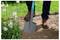 Une personne portant des bottes brunes qui creuse la terre avec une pelle grise près de fleurs blanches dans un jardin, suggérant des activités de jardinage ou de plantation.