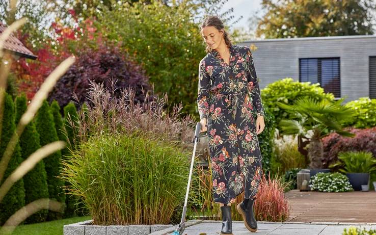 Une femme en robe fleurie arrose des plantes sur une terrasse, entourée de végétation verte et rouge dans un jardin par temps ensoleillé.