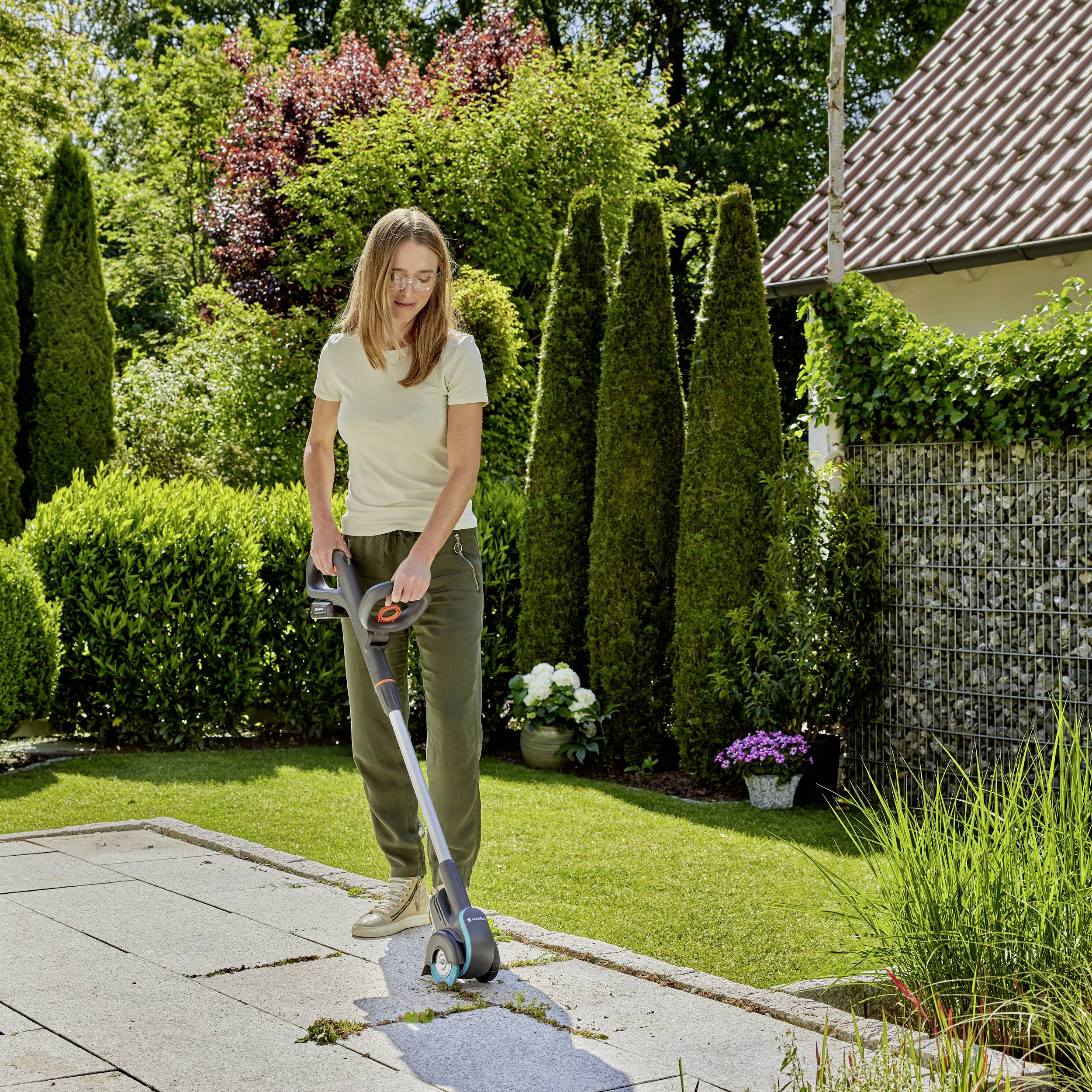 Une femme tond la pelouse dans un jardin ensoleillé à l'aide d'une tondeuse électrique. Des arbres et une maison sont visibles en arrière-plan.