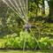 Un arroseur de jardin diffuse des jets d'eau sur une pelouse bien entretenue, entourée de buissons, sous un ciel ensoleillé.