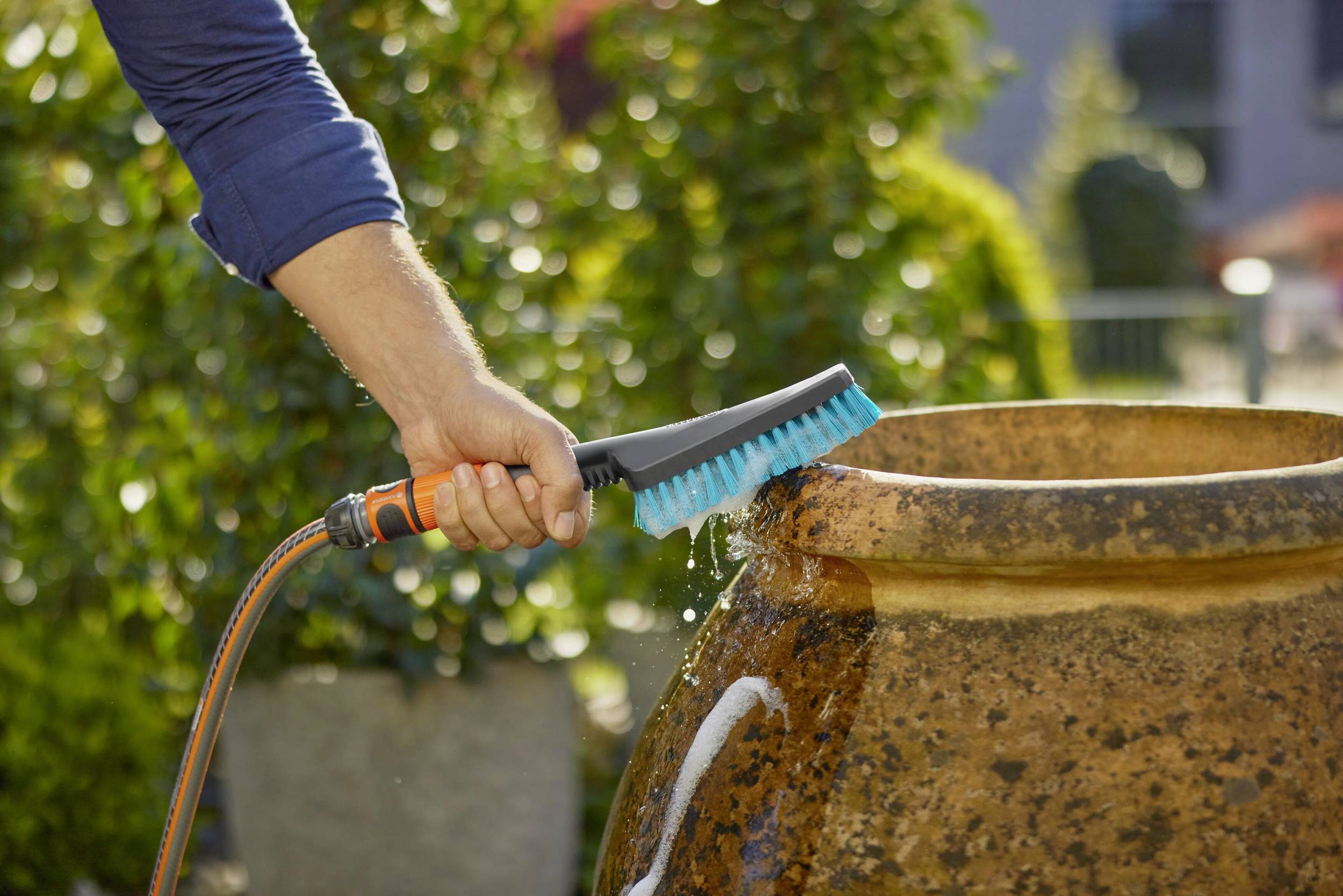 Une personne nettoie un grand pot en céramique rond à l'extérieur avec une brosse à eau. L'arrière-plan est flou avec des buissons.