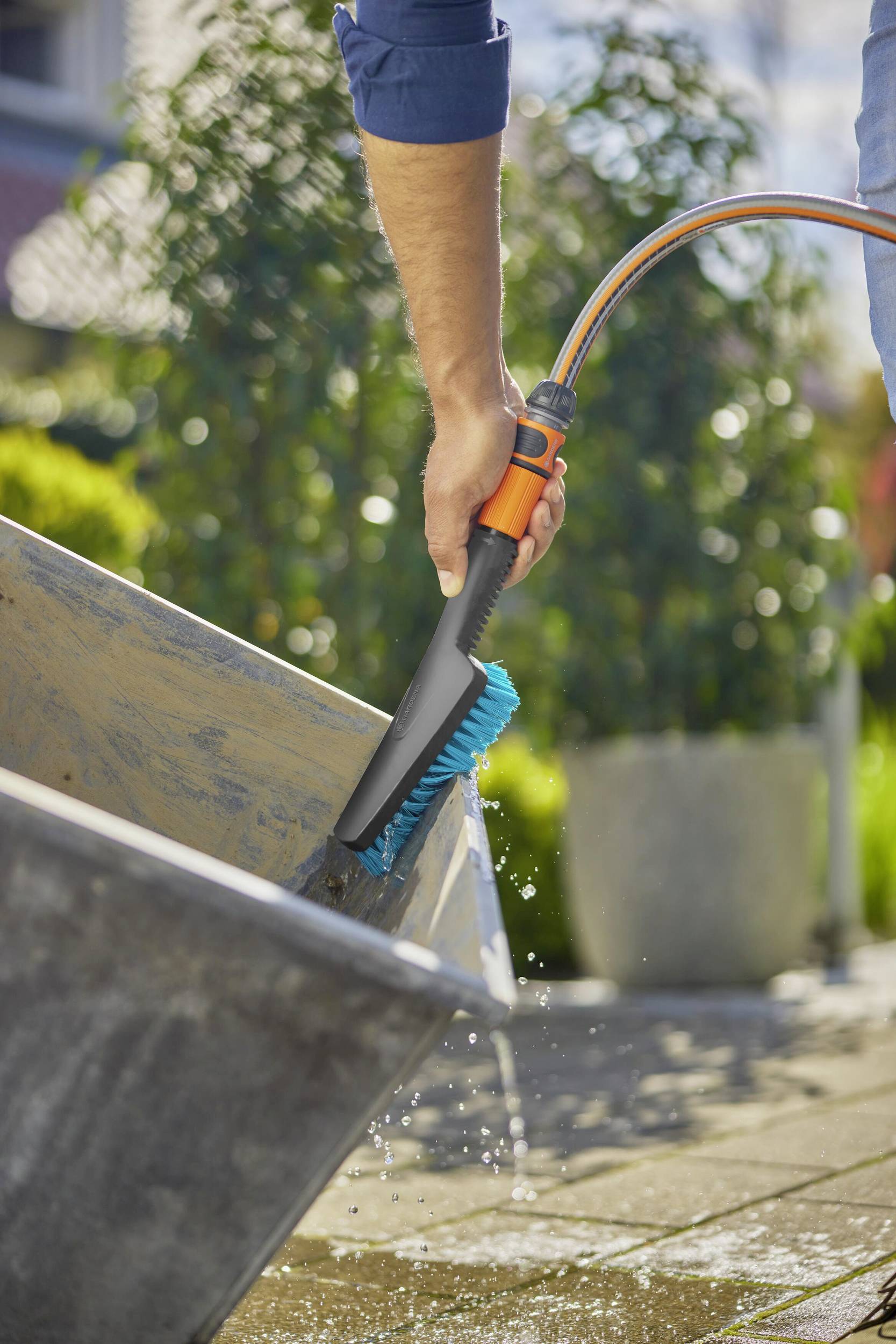 Une personne utilise une brosse à eau pour nettoyer un seau à l'extérieur. Le seau est placé sur un sol pavé devant des plantes.