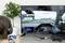 Une personne joue à un jeu de course automobile sur un téléviseur, tenant une manette bleue à la main. Un arbre se trouve en arrière-plan.