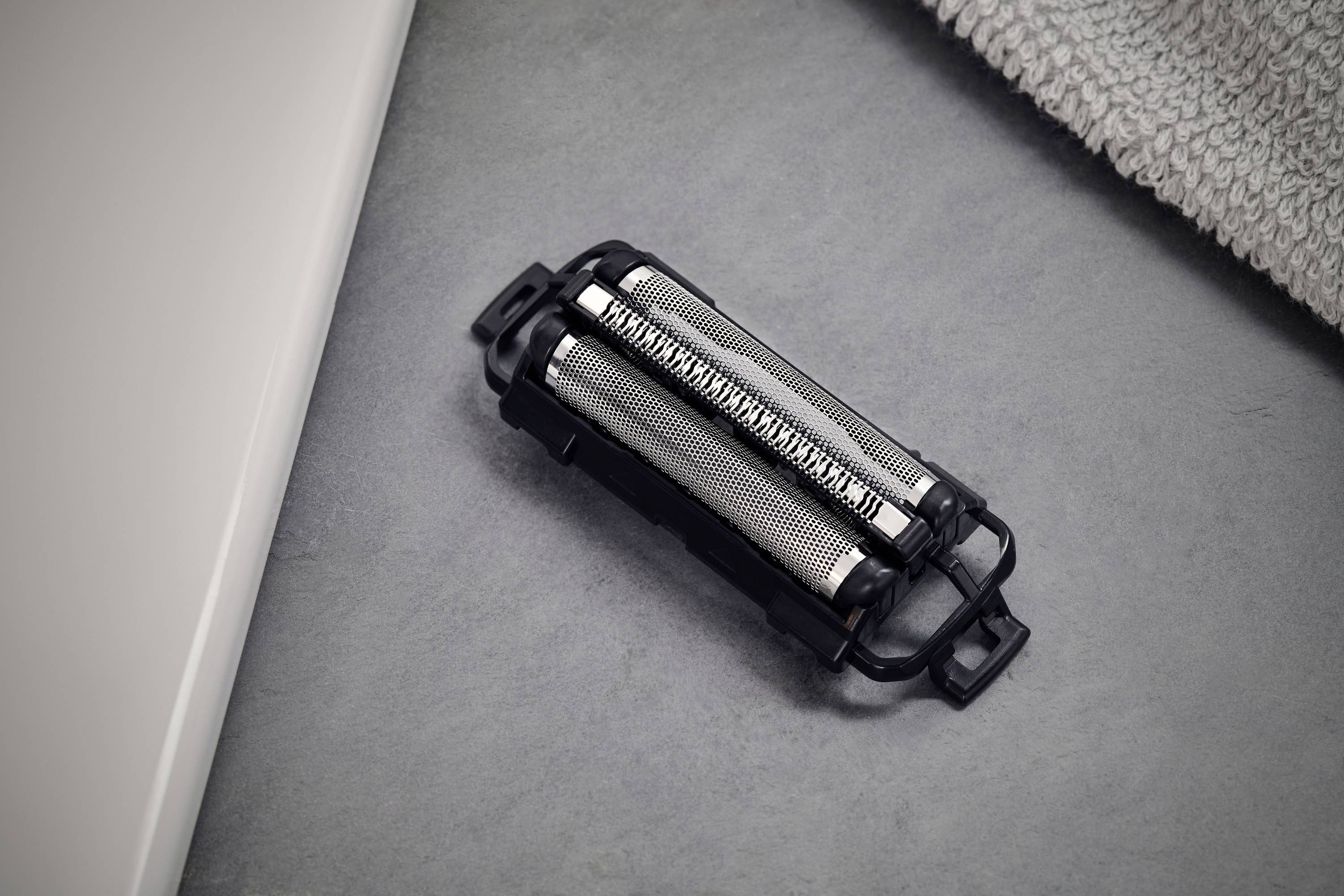 Tête de rasoir sur un fond gris, à côté d'une serviette et du bord d'un lavabo blanc.