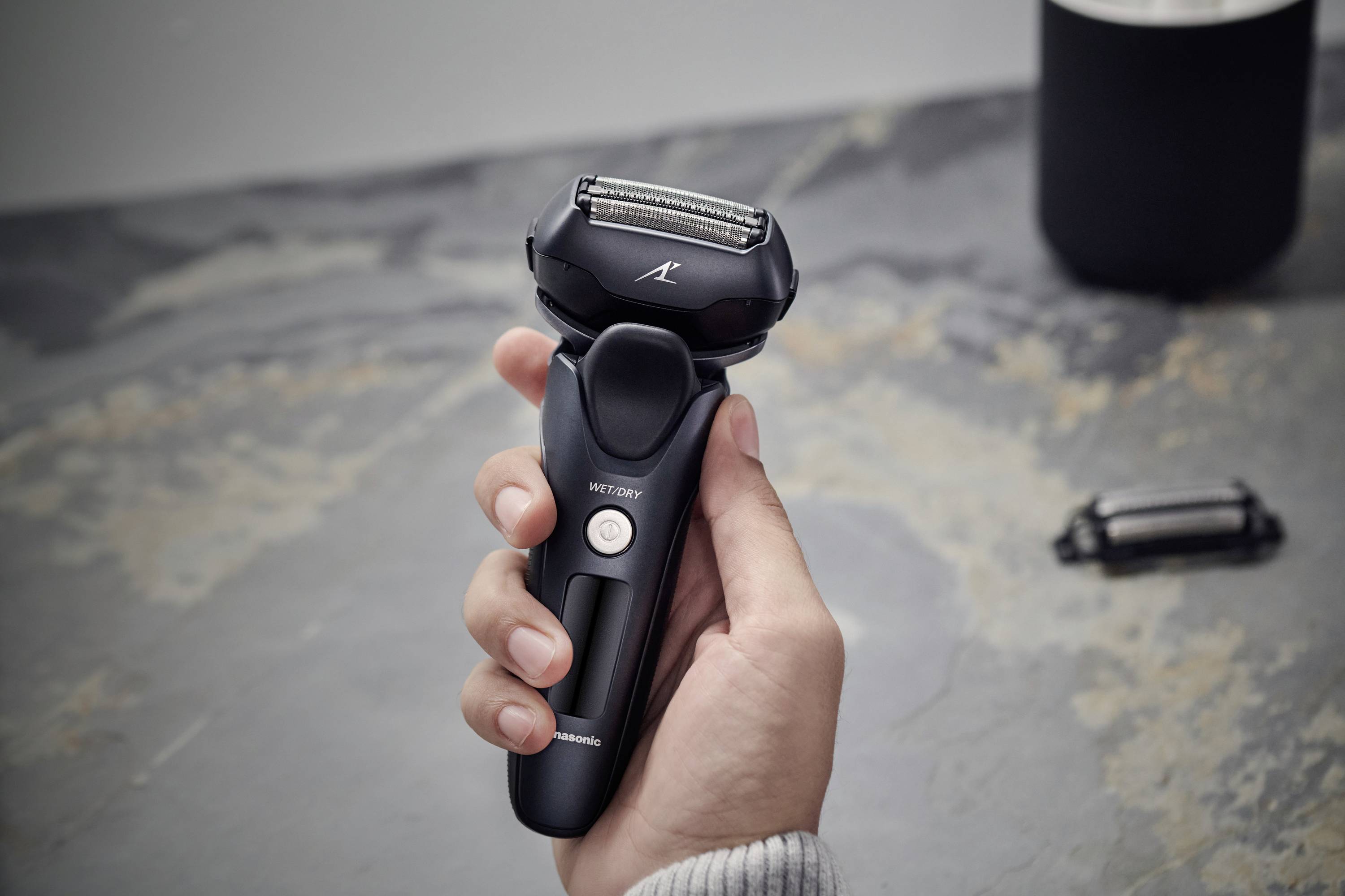 Une main tient un rasoir électrique moderne au-dessus d'une surface grise. À droite en arrière-plan se trouve une pièce détachée du rasoir.