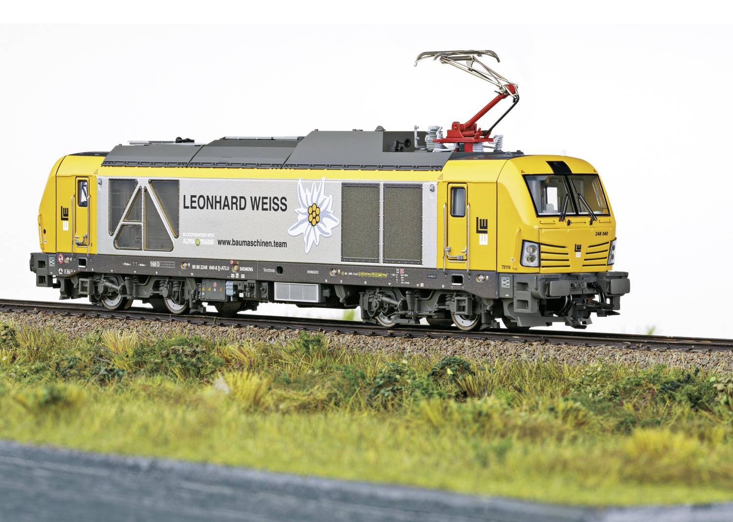 Locomotive jaune avec une bande grise et le logo 'Leonhard Weiss' circulant sur un rail, dans un environnement herbeux avec une voie ferrée.