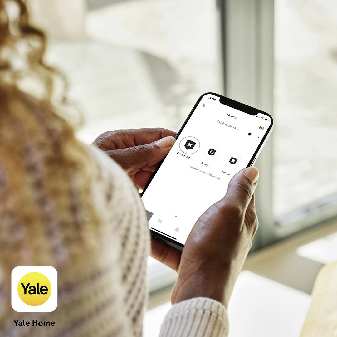 Une personne tient un smartphone avec l'application 'Yale Home' ouverte. L'application affiche des options de domotique.