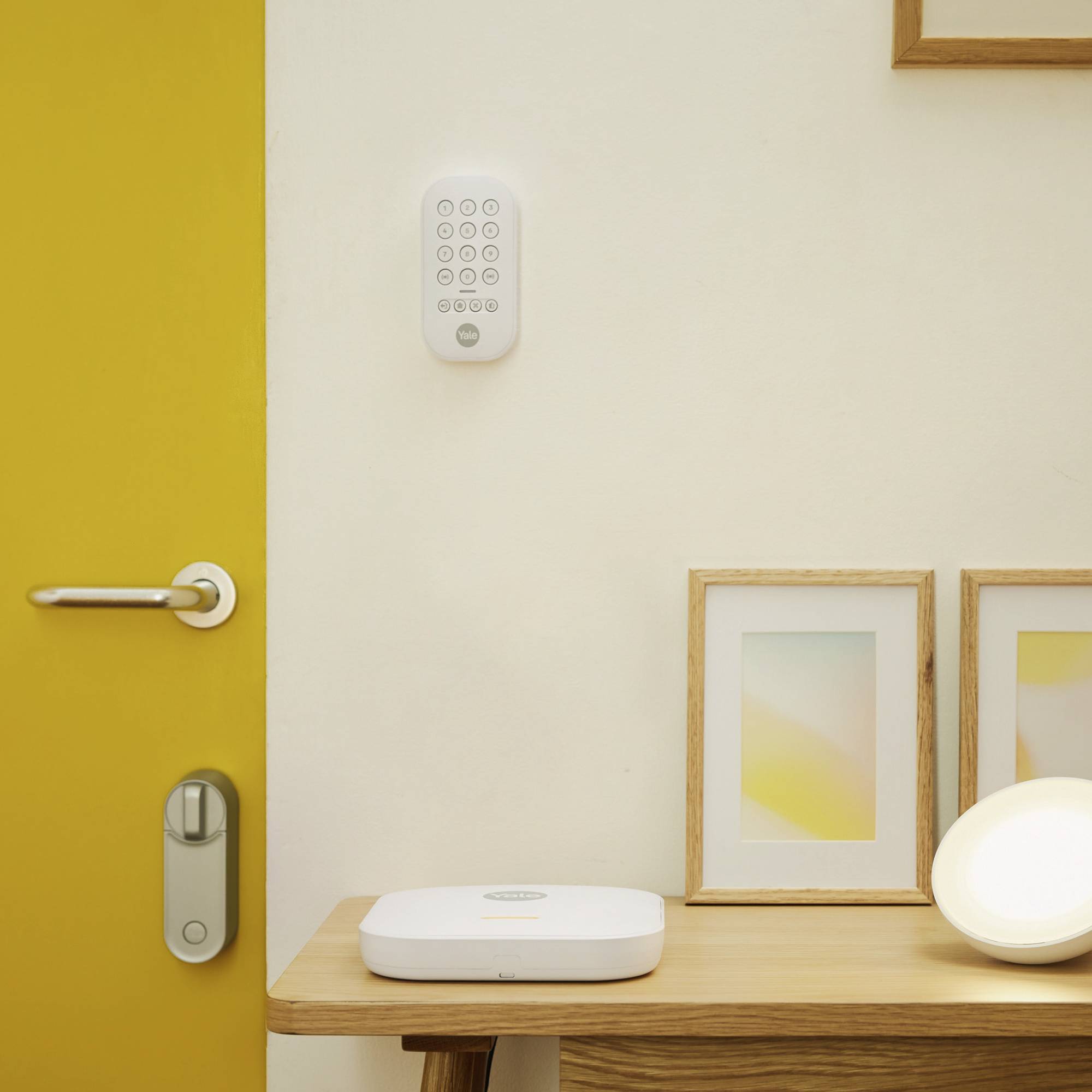 Une porte jaune avec une serrure numérique, à côté d'un mur équipé d'un clavier. En dessous, une table supportant deux cadres photo et une boîte blanche.