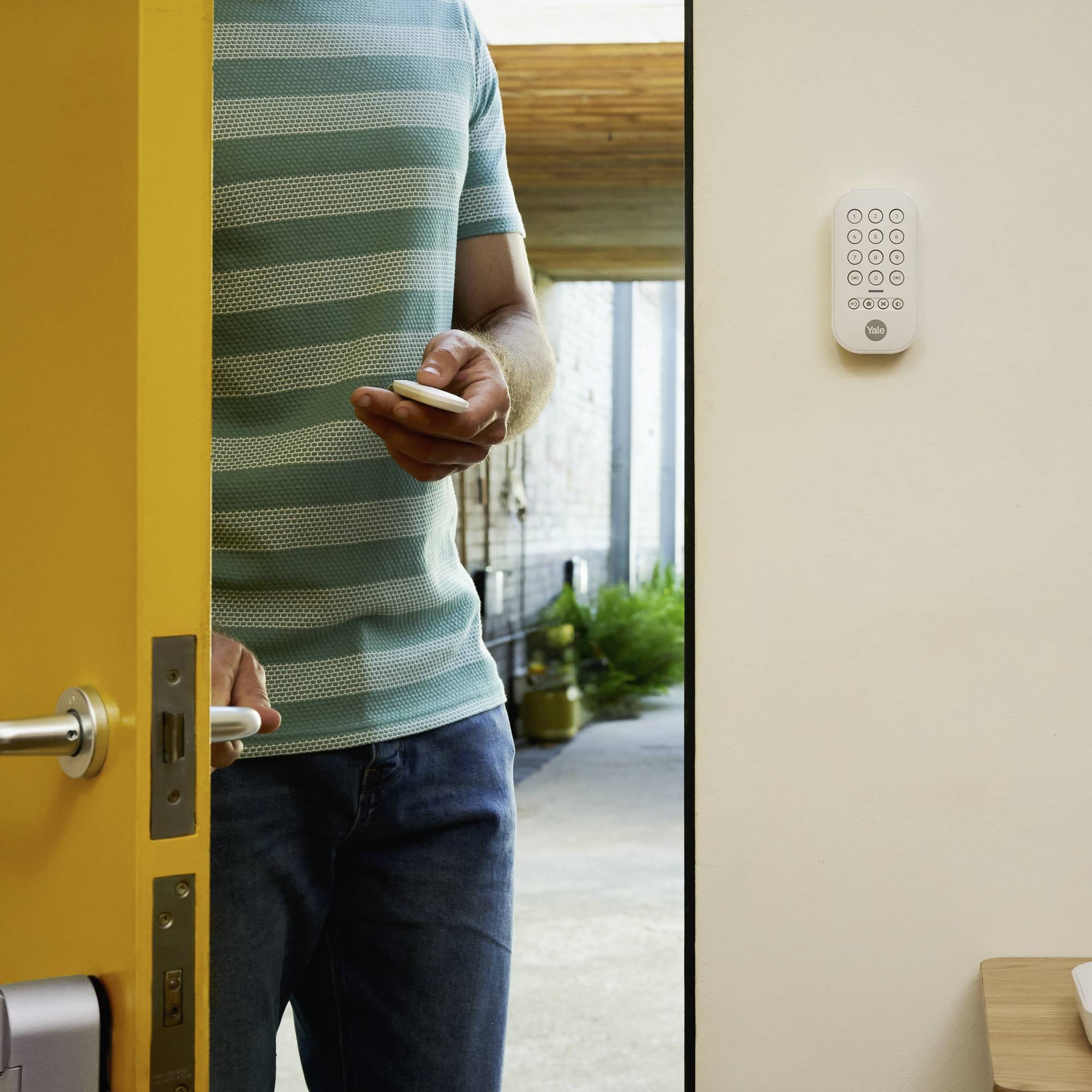 Un homme portant un t-shirt rayé ouvre une porte jaune, tenant un smartphone à la main, et se tient partiellement visible dans l'embrasure de la porte.