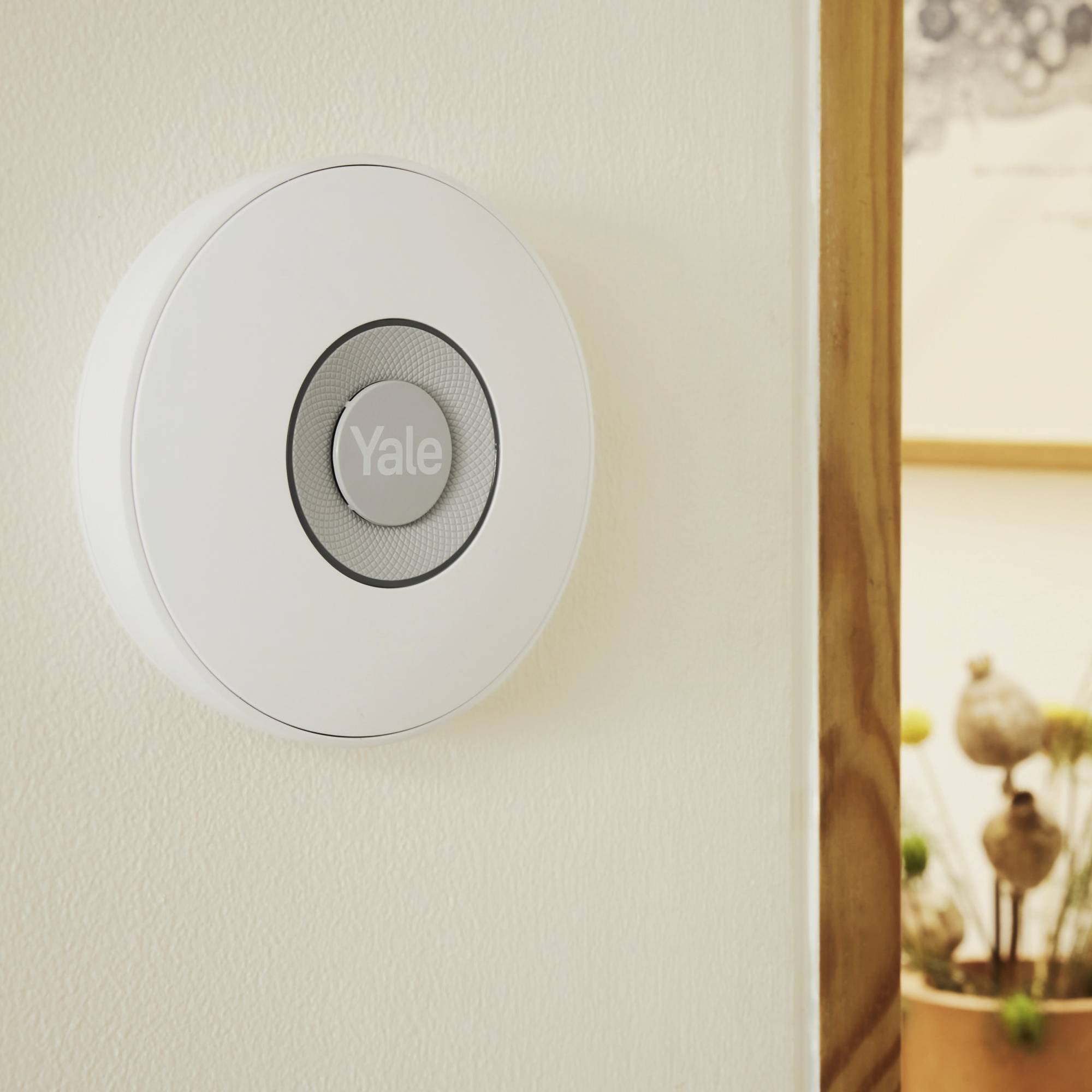 Une serrure ronde blanche avec 'Yale' au centre, montée sur un mur à côté d'une porte en bois. Une plante décorative est visible en arrière-plan.