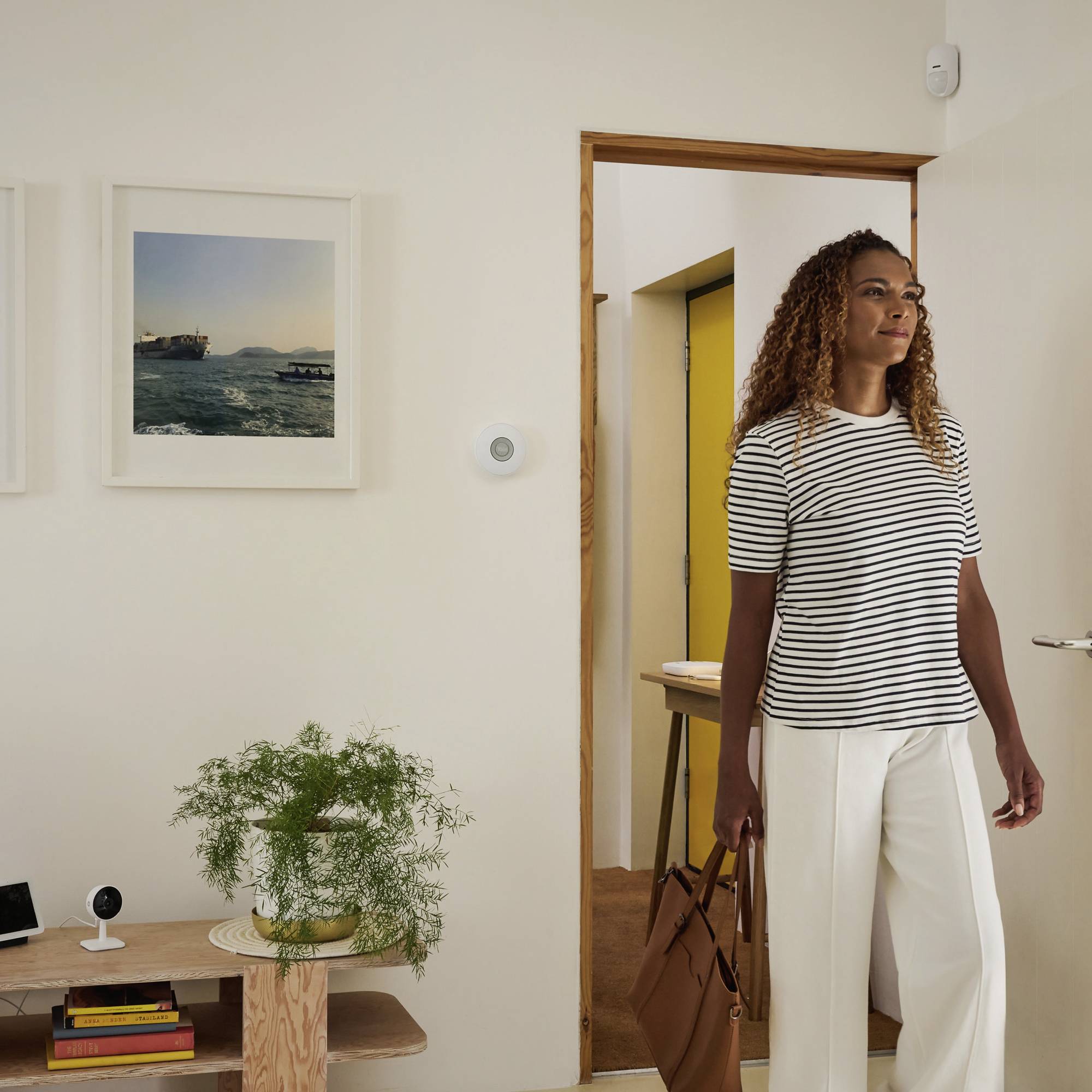 Une femme aux cheveux bouclés porte un chemisier rayé et un pantalon blanc, et se tient dans une pièce avec des tableaux aux murs et une petite table.
