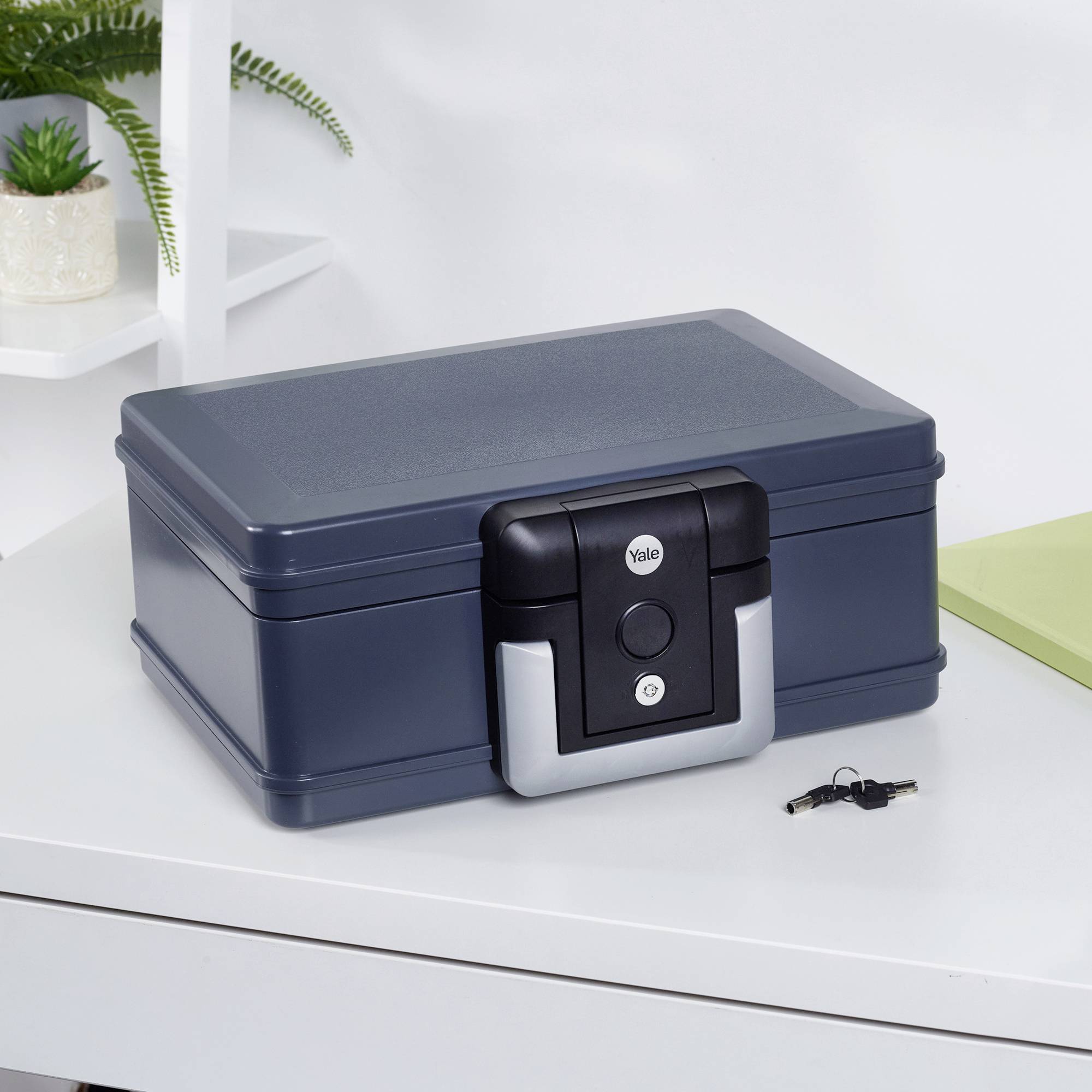 Un coffre-fort bleu-gris sur un bureau avec des plantes en arrière-plan. Des clés sont posées à côté.