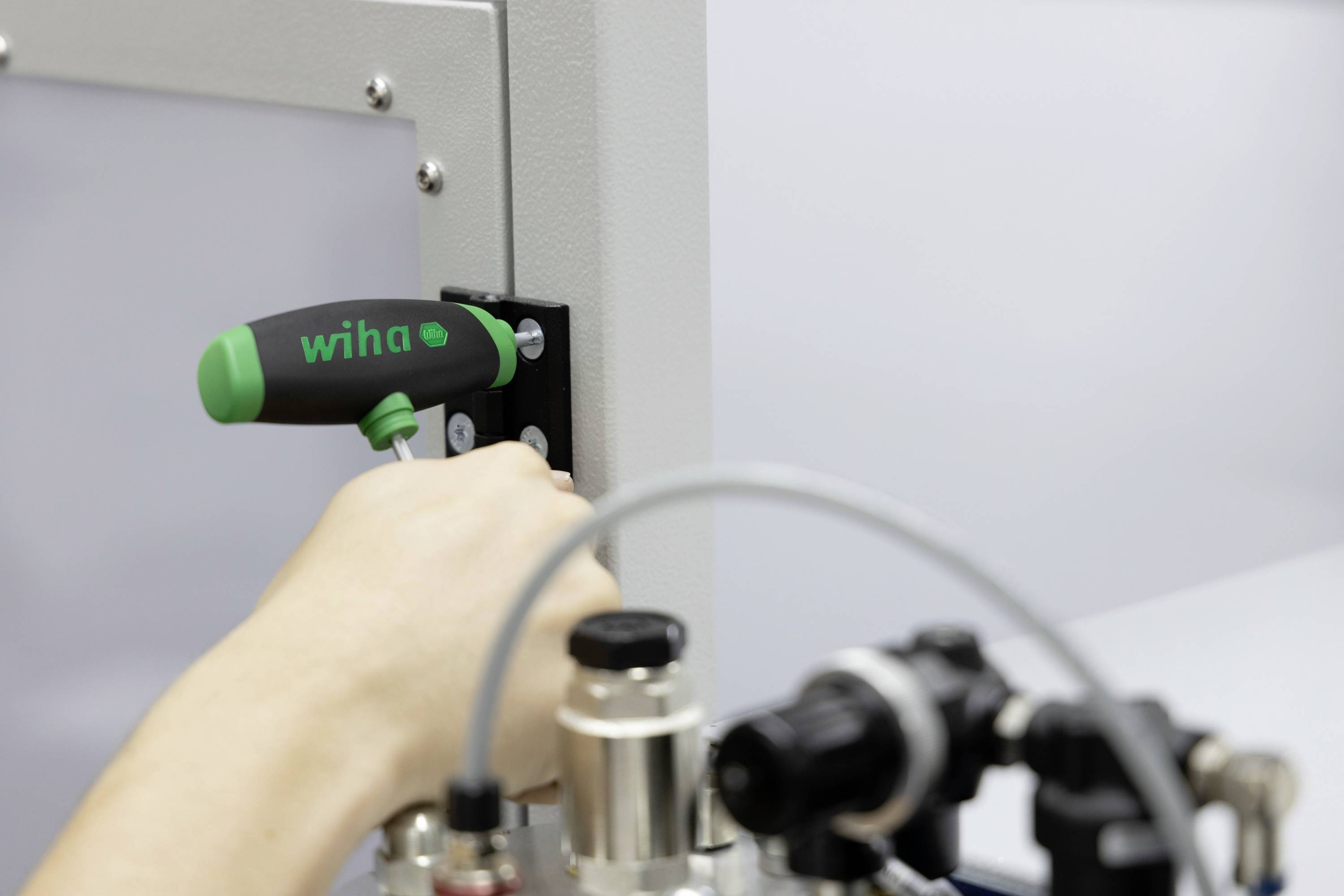 Une main utilise un tournevis vert pour visser une vis sur une grande machine grise. Des tuyaux et des vannes sont visibles au premier plan.