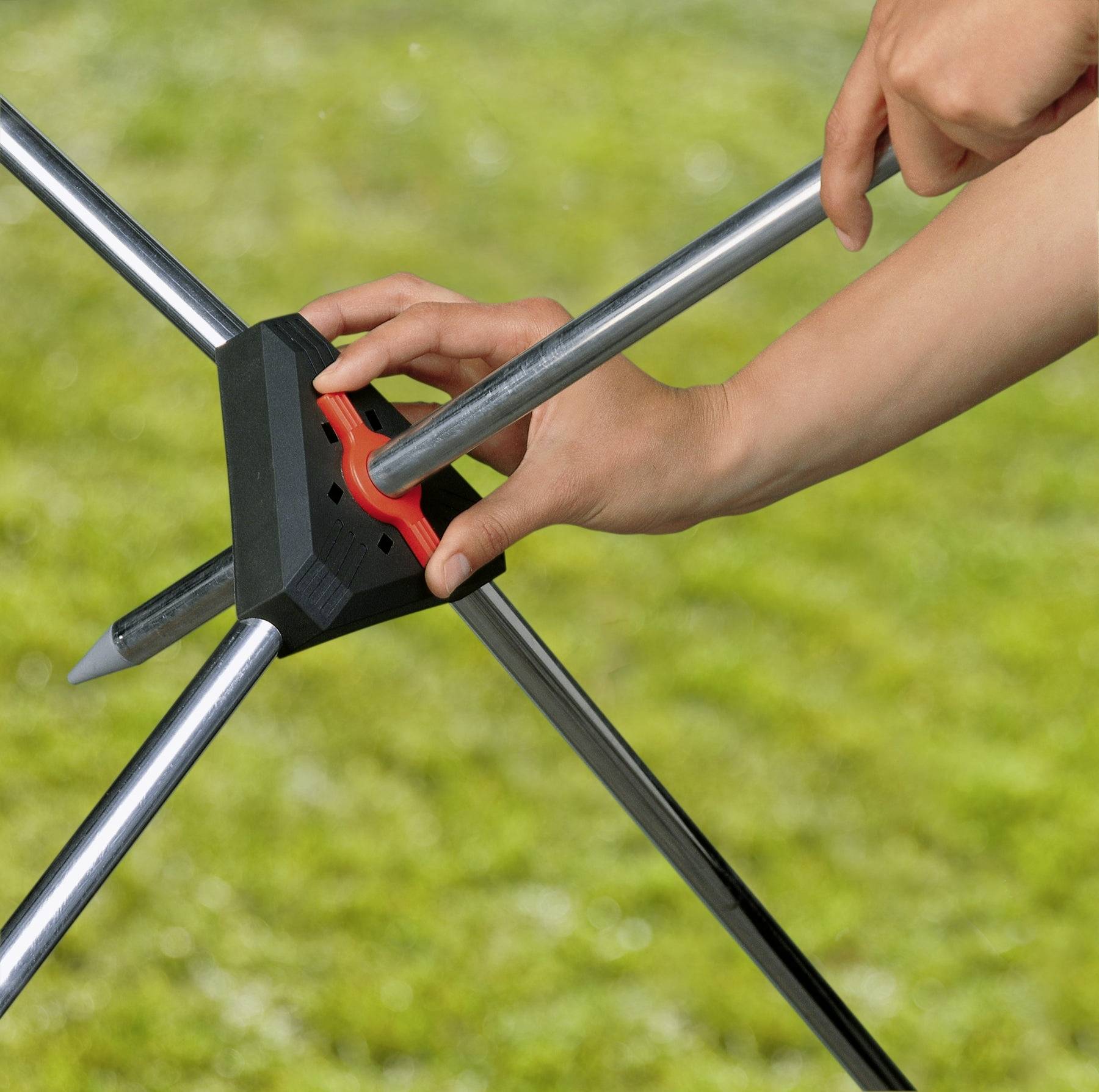 Une personne utilise un outil rouge pour ajuster l'angle d'un trépied métallique sur l'herbe, en se concentrant sur le mécanisme de verrouillage de ses pieds.