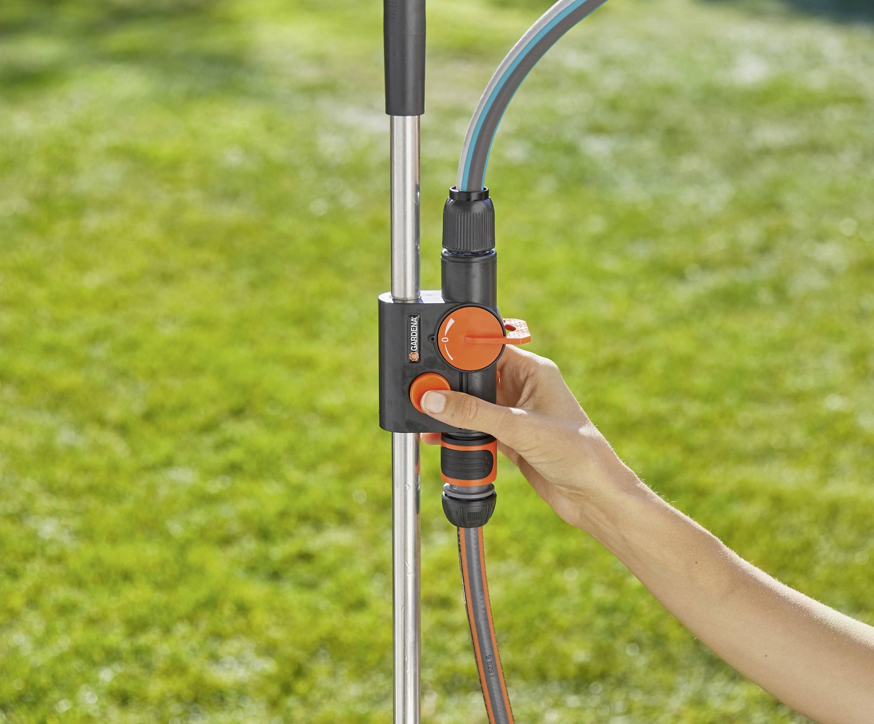 Une main ajuste une vanne de contrôle du débit d'eau de jardin noir et orange fixée à un poteau métallique, avec une pelouse visible en arrière-plan.