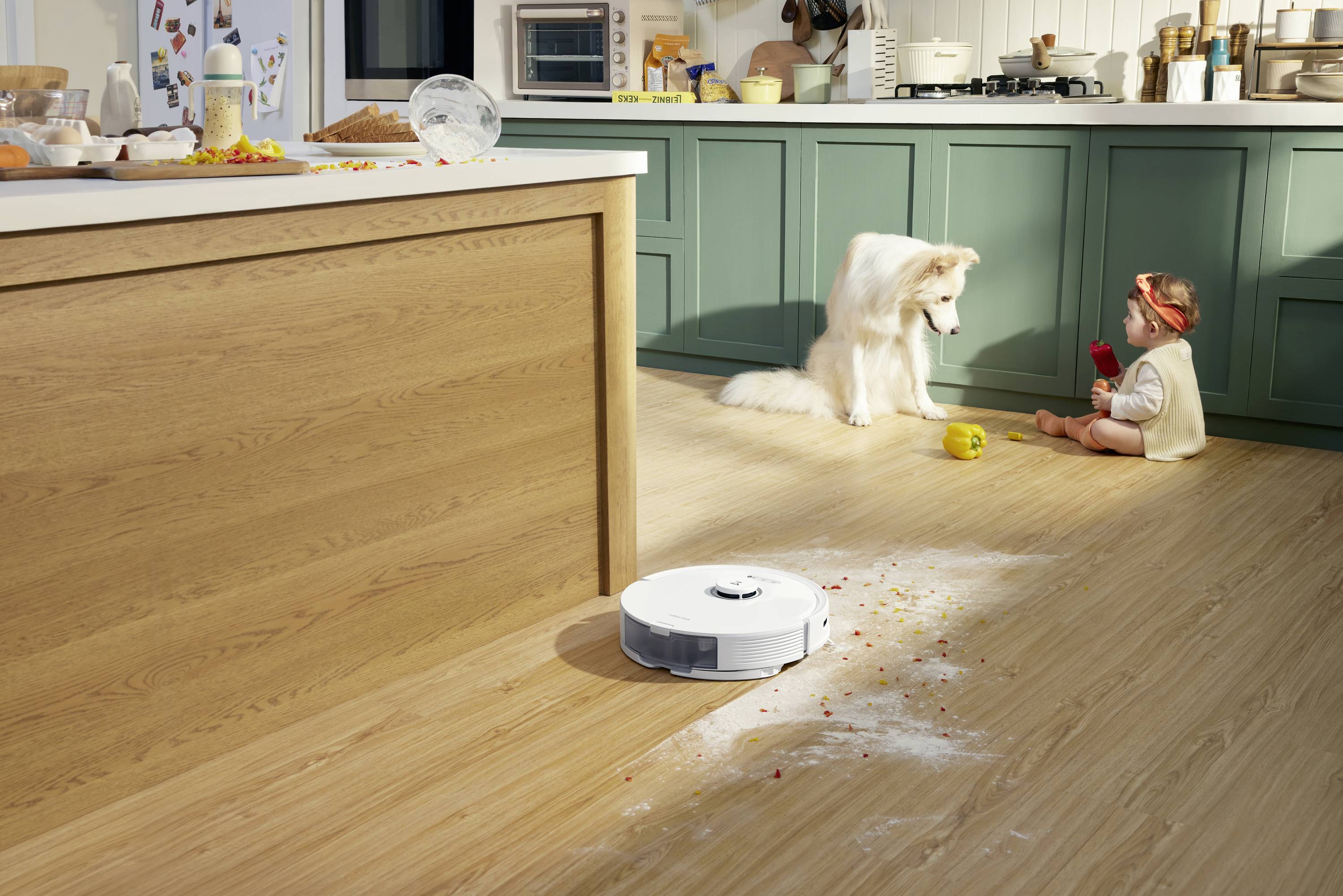 Un robot ménager nettoie de la farine sur un plancher en bois. En arrière-plan, un jeune enfant joue avec un chien dans la cuisine.