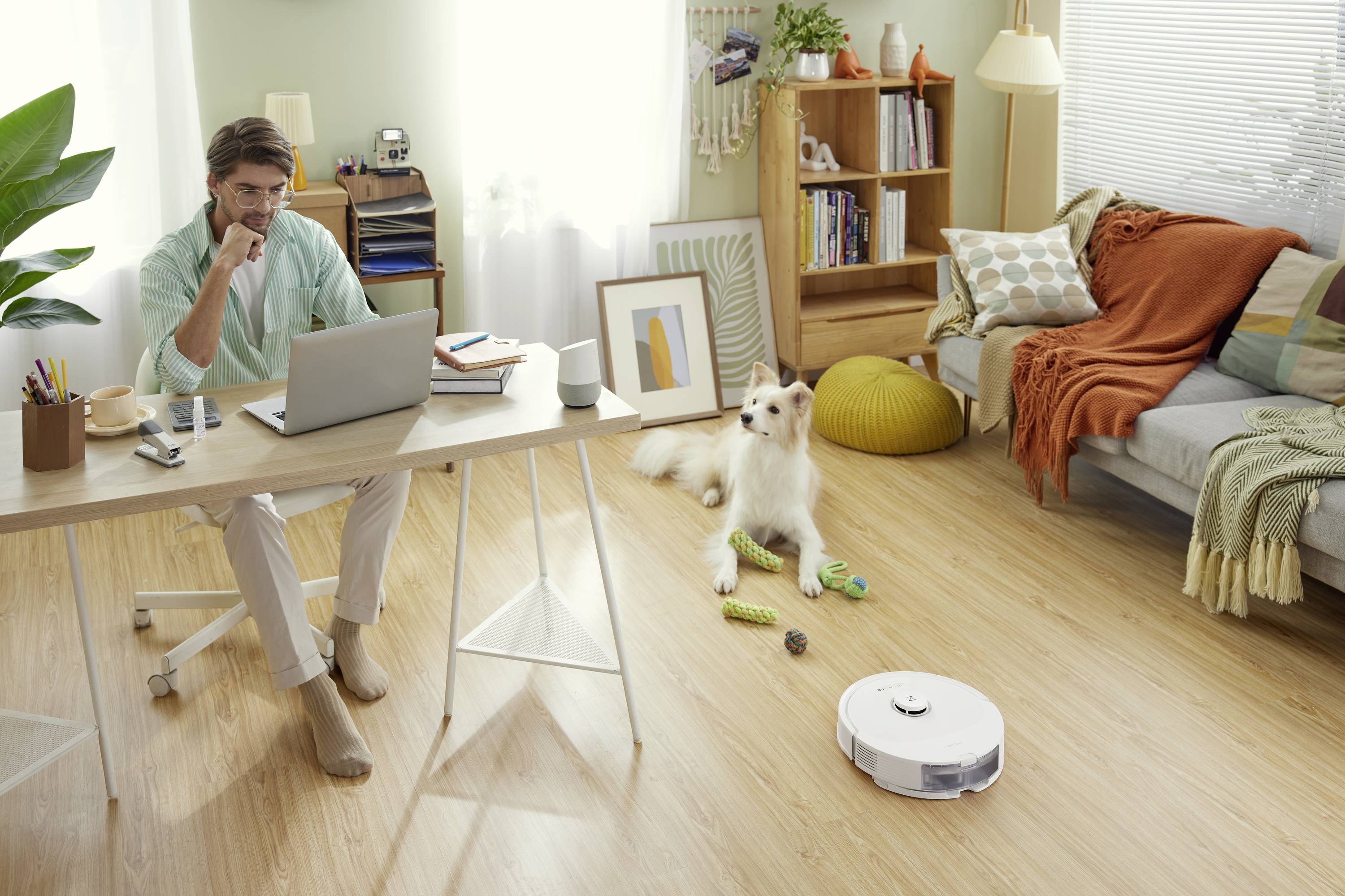 Un homme travaille sur un ordinateur portable dans un salon confortable. Un chien est allongé à côté de lui, tandis qu'un aspirateur robot nettoie le sol.