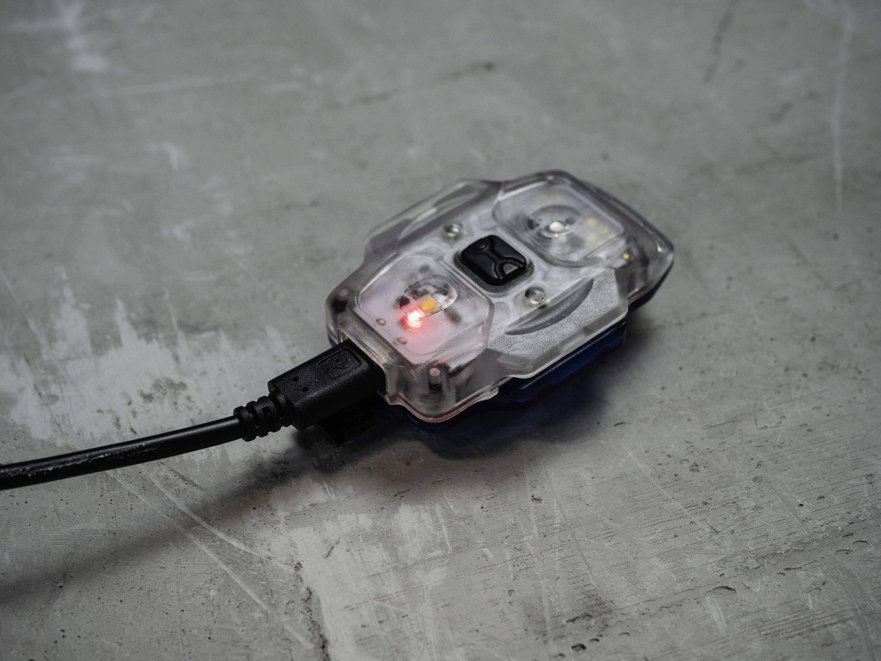 Un dispositif en plastique transparent sur un sol en béton, avec une lumière rouge vive au centre, relié par un câble USB sur le côté gauche.
