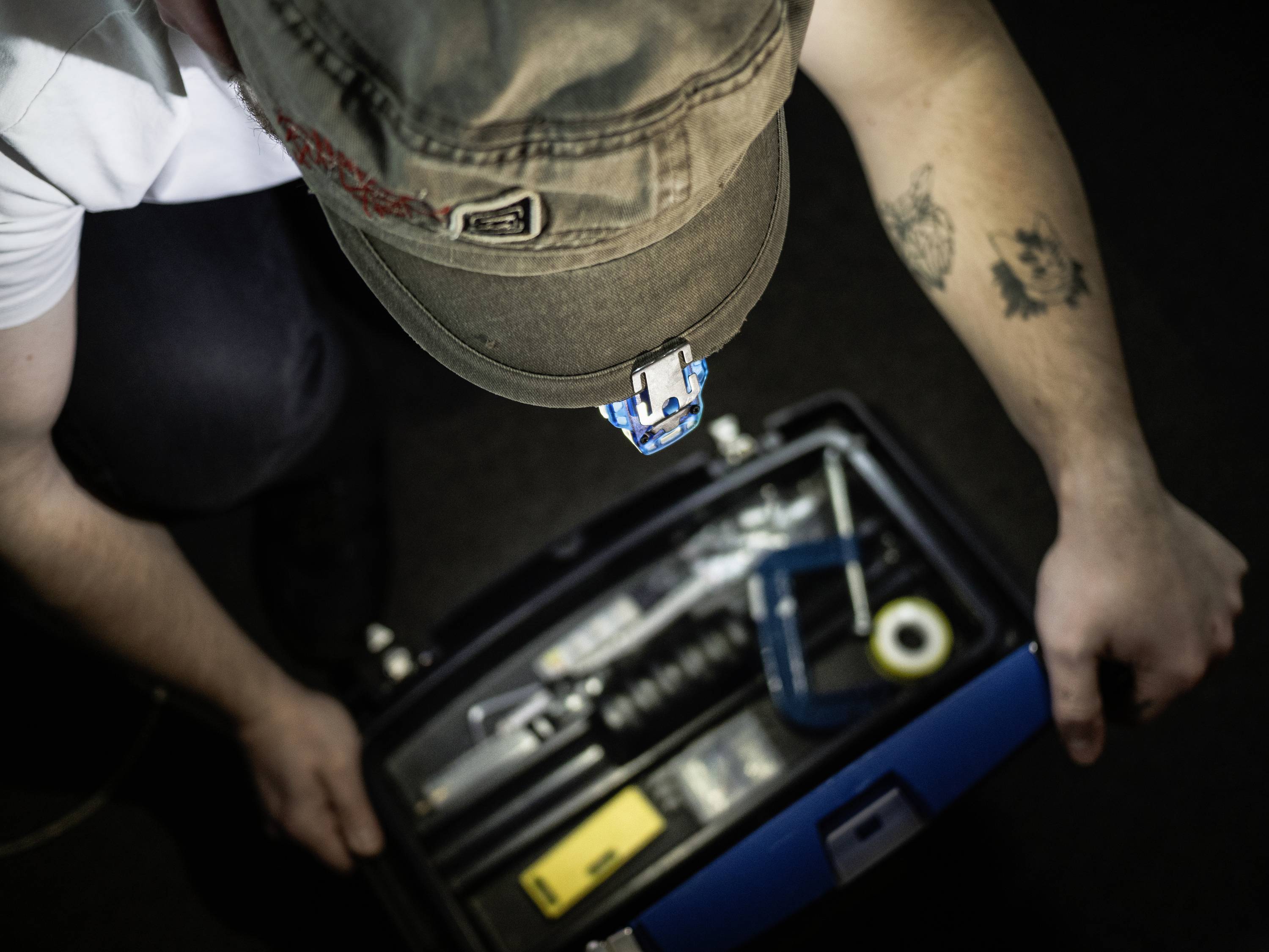 Une personne ouvre une boîte à outils. Vue de dessus, divers outils sont visibles. La personne porte une casquette et a un tatouage sur le bras.
