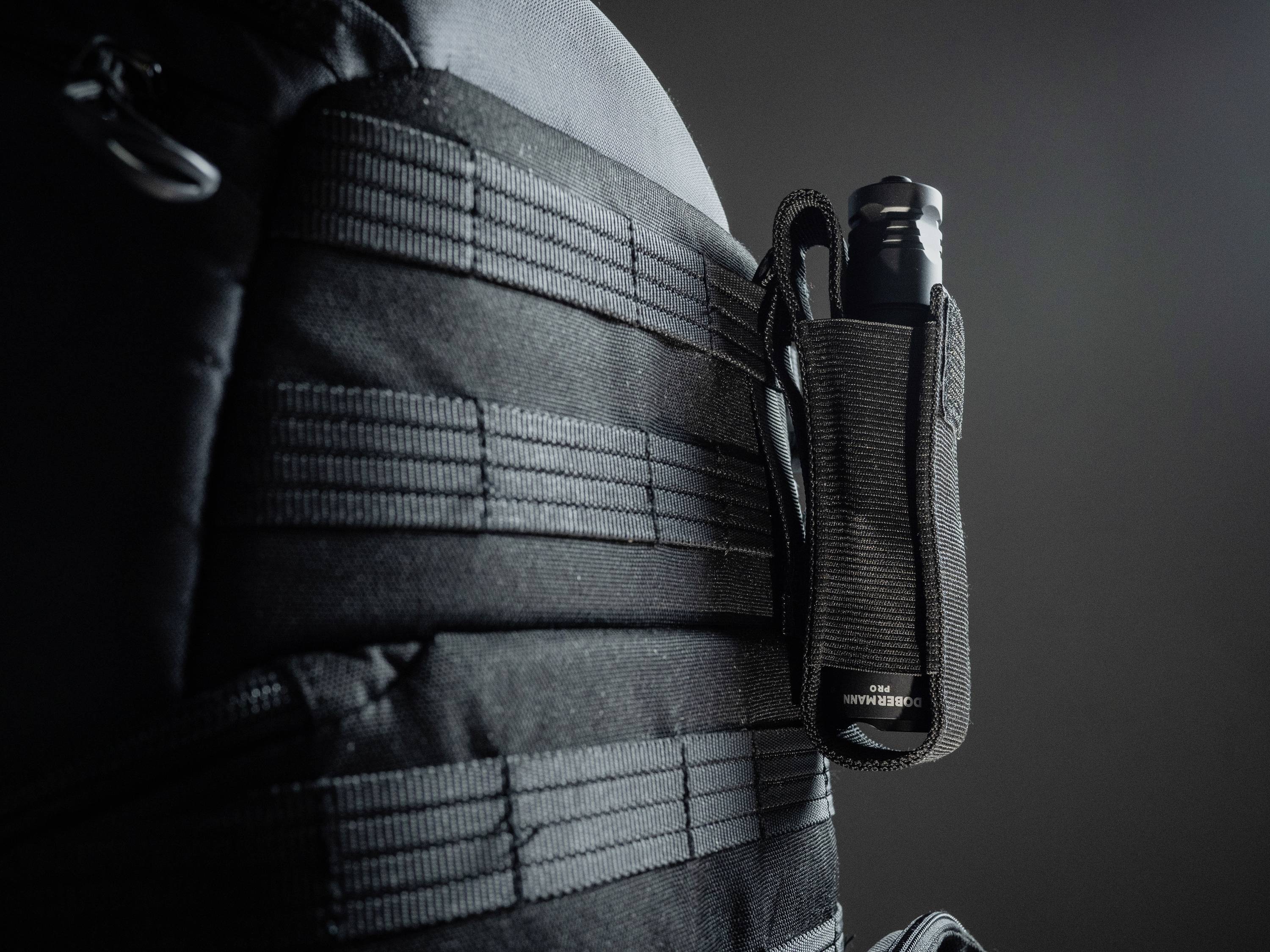 Un sac tactique noir avec des sangles MOLLE. Un étui pour lampe tactique est fixé sur le côté.