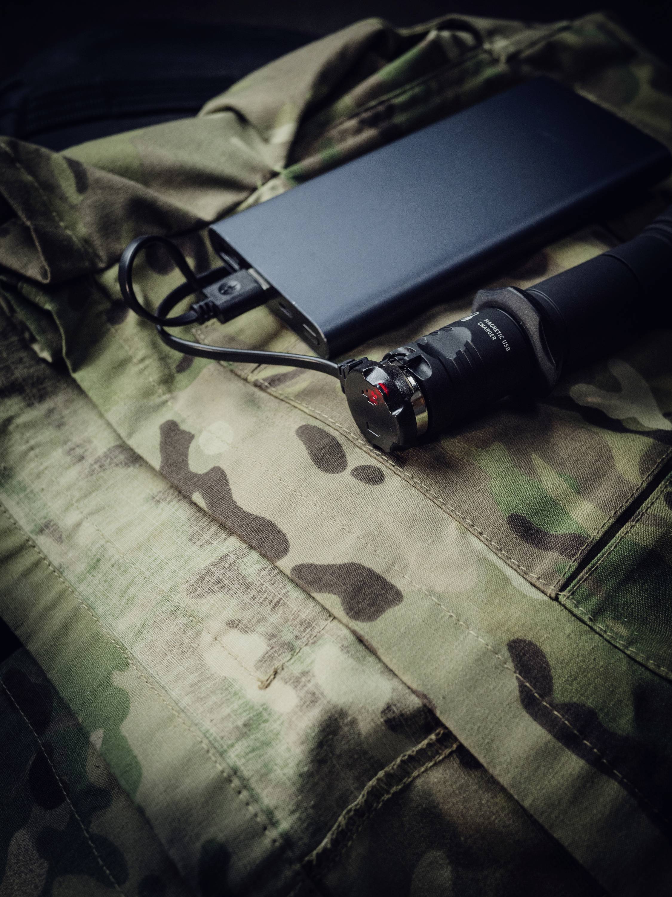 Une batterie externe noire est posée sur une veste de camouflage, à côté d'un module de lampe tactique relié par un câble.