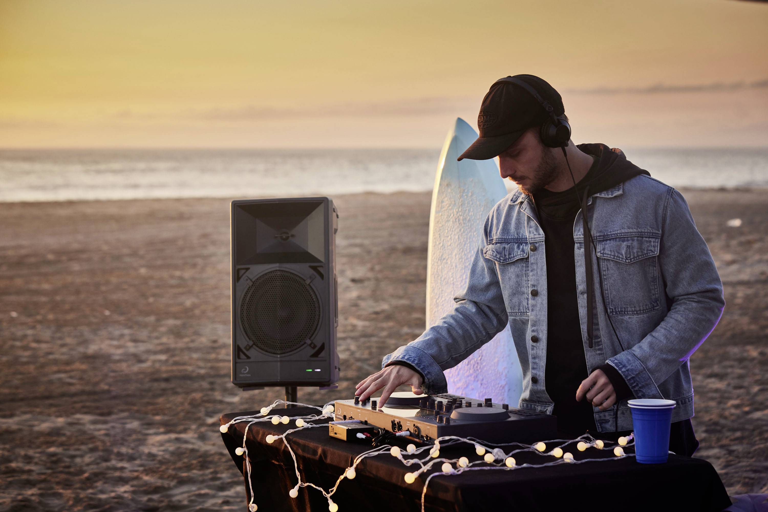 Une personne portant des écouteurs diffuse de la musique sur une plage au coucher du soleil. Au premier plan, se trouve une table avec un équipement de DJ et des lumières.