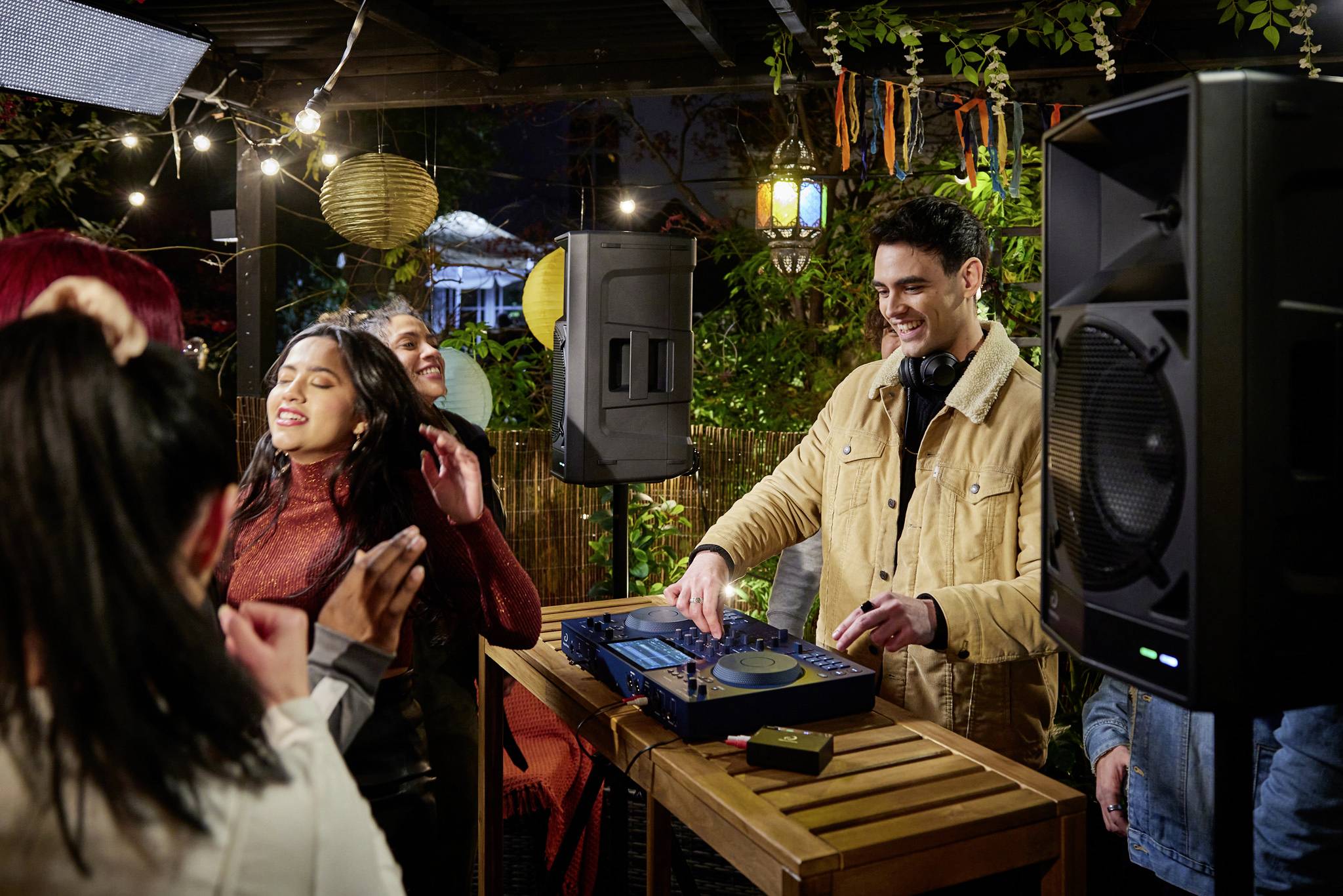 Un groupe de personnes danse et sourit pendant qu'un DJ passe de la musique, entouré de lumières colorées dans un cadre extérieur chaleureux.