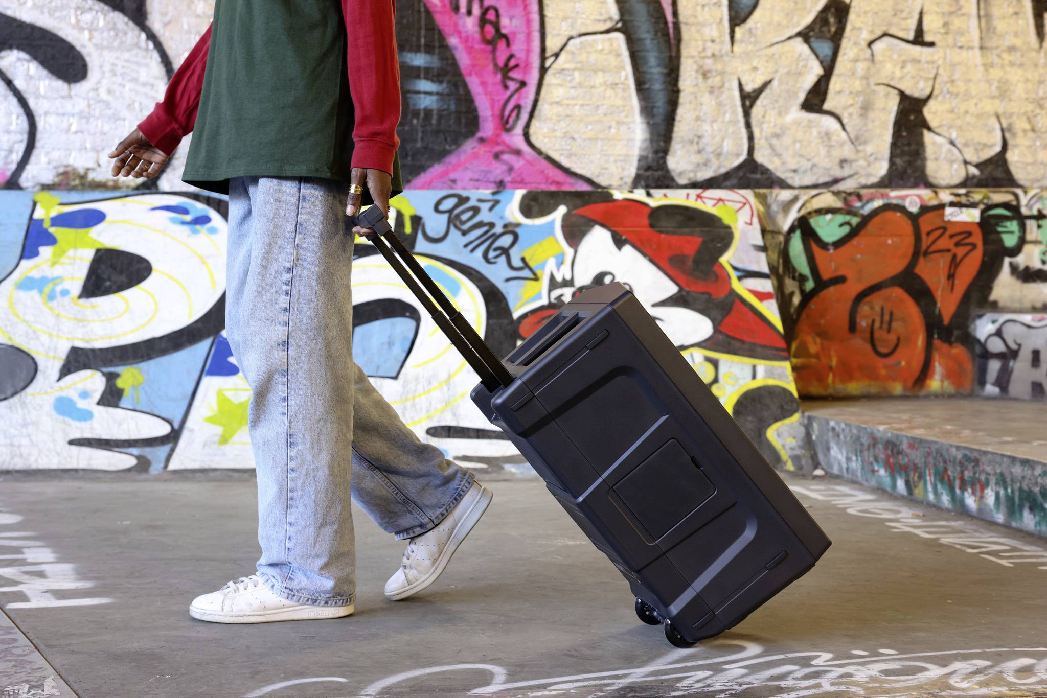 Une personne tire une valise noire à roulettes dans un environnement urbain aux murs ornés de graffitis colorés.